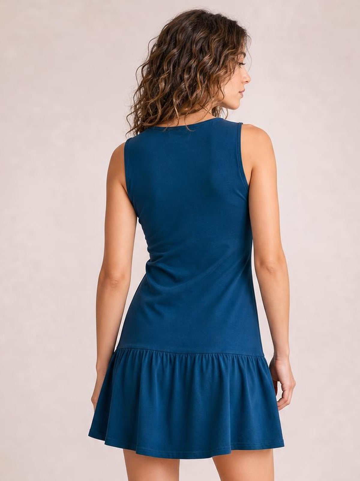 A plus-size model stands facing away in a deep blue sleeveless mini dress with a fitted bodice, gathered waist seam, and gently flared skirt.