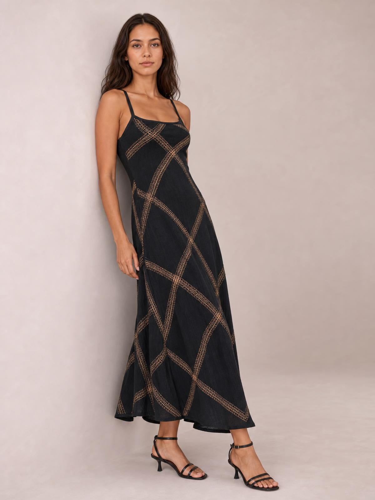 Model wears a black square-neck maxi dress with thin straps, a fitted bodice, flowing flared skirt, and tan abstract line pattern.