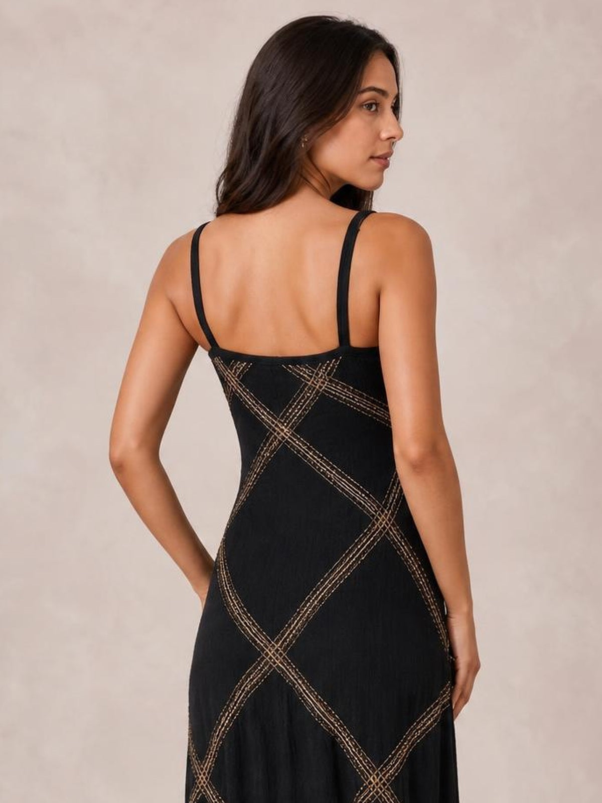 A model poses from the back in a black maxi dress with thin shoulder straps, a fitted bodice, and contrasting geometric stitched detailing.