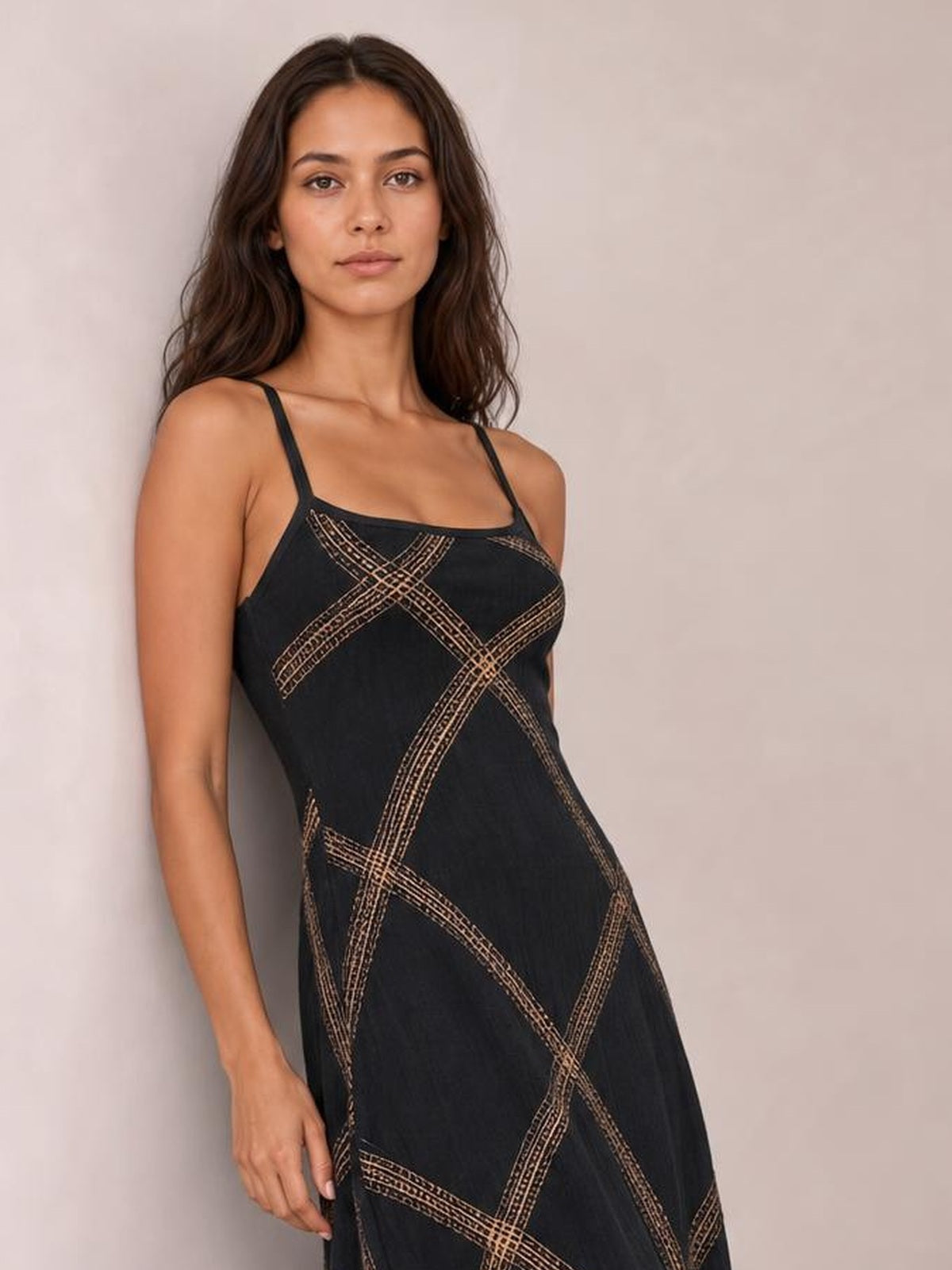 A model leans in a black square-neck maxi dress with thin shoulder straps, a fitted bodice, and a flowing silhouette patterned with intersecting tan lines.