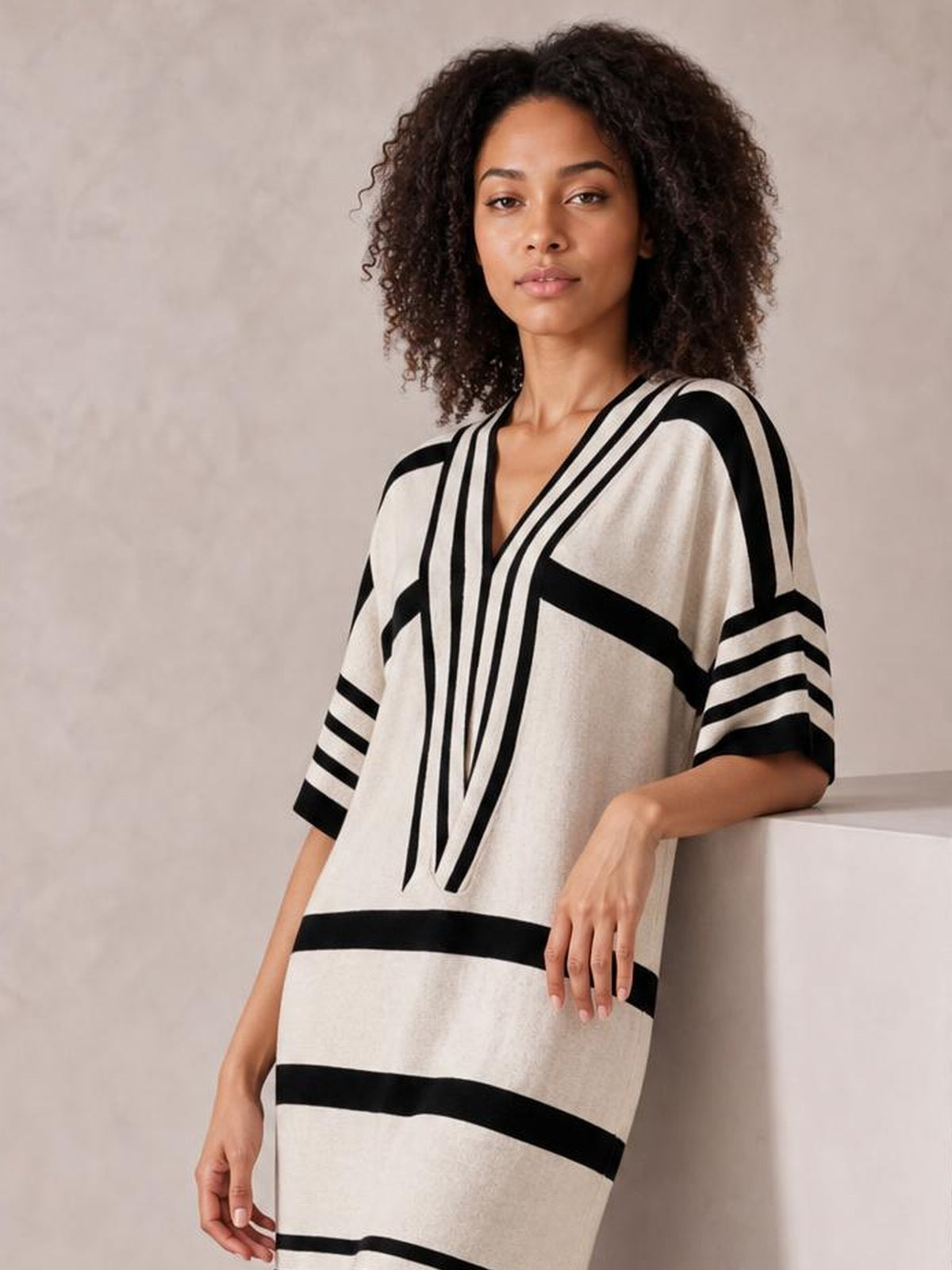 Model leans against a ledge wearing a cream and black striped half-sleeve midi dress with a V-neckline and relaxed straight silhouette.