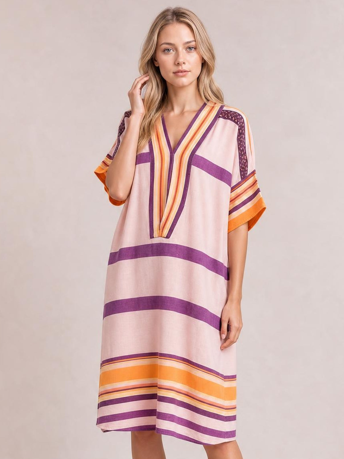 Model poses in a loose striped half-sleeve midi dress with a V-neck, pale pink base, purple and orange bands, and a relaxed straight silhouette.