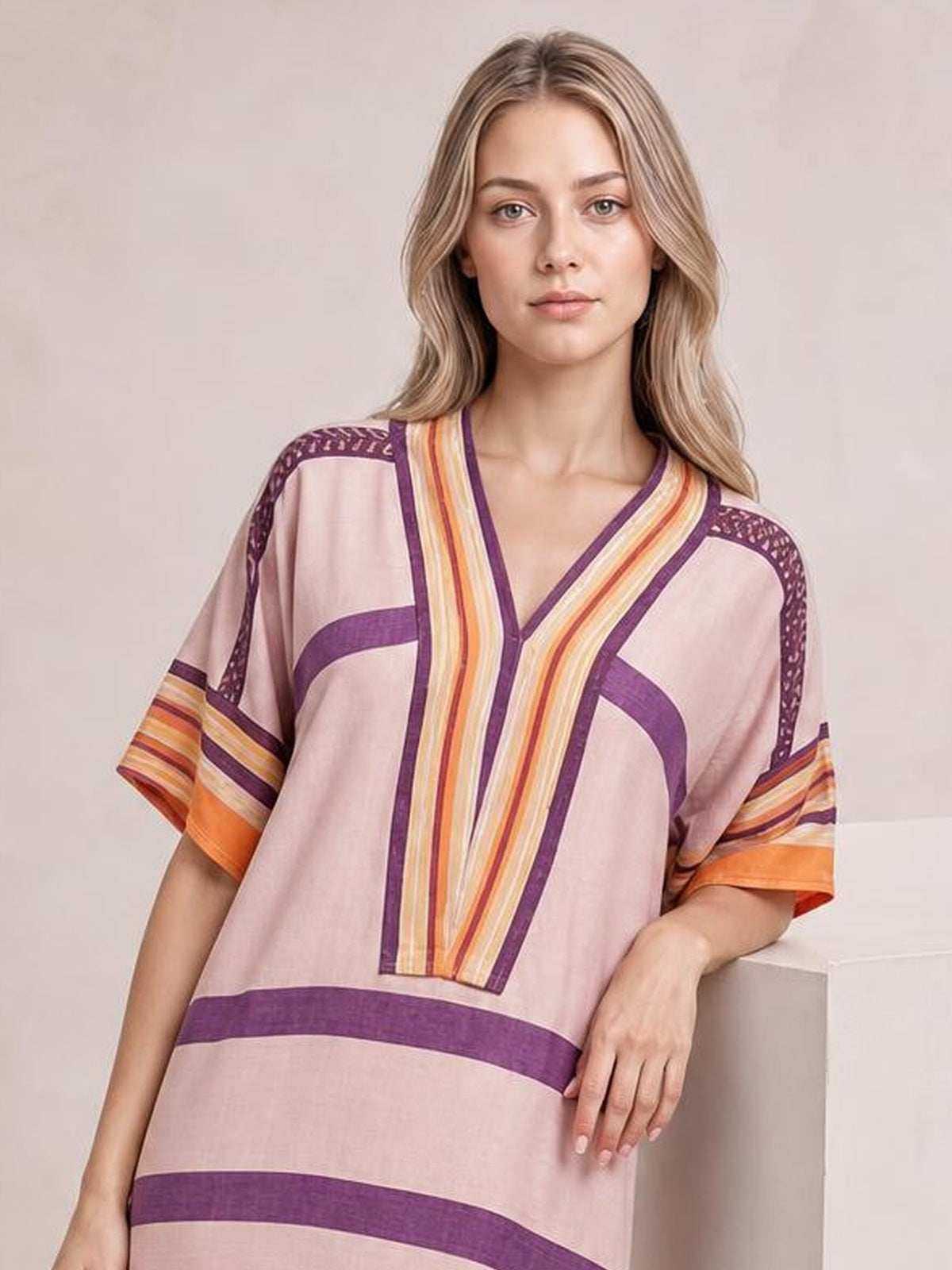 A model wears a loose pink striped midi dress with half sleeves, a V-neckline, bold purple and orange trim, and a relaxed silhouette.