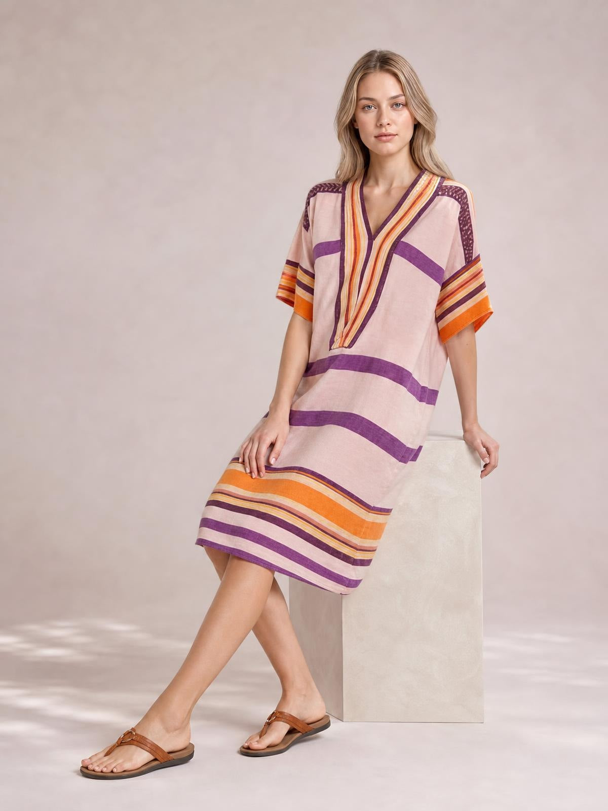 A model poses with crossed legs in a loose pink striped midi dress featuring half sleeves, a V-neck, purple and orange bands, and a straight silhouette.