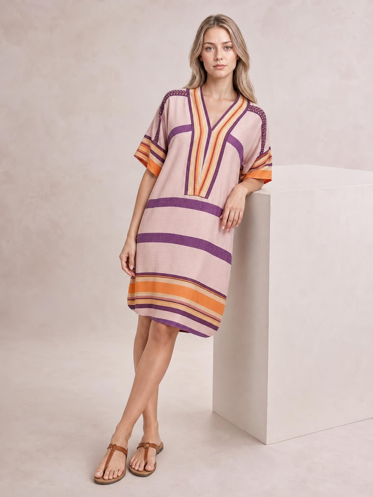 A model leans against a pedestal in a pink striped midi dress with half sleeves, a V-neck, colorful trim, and a relaxed straight silhouette.