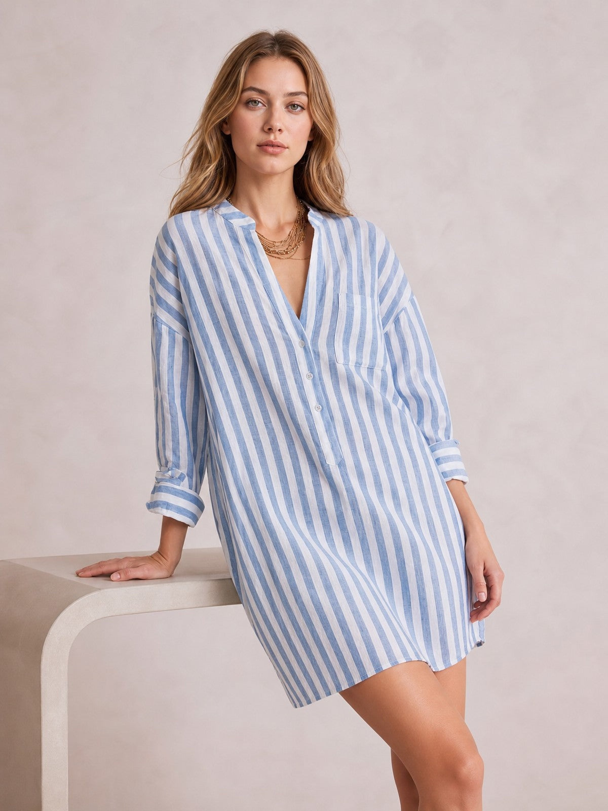 A model leans against a table in a blue-and-white striped shirt mini dress with a relaxed silhouette, button front, chest pocket, and rolled long sleeves.