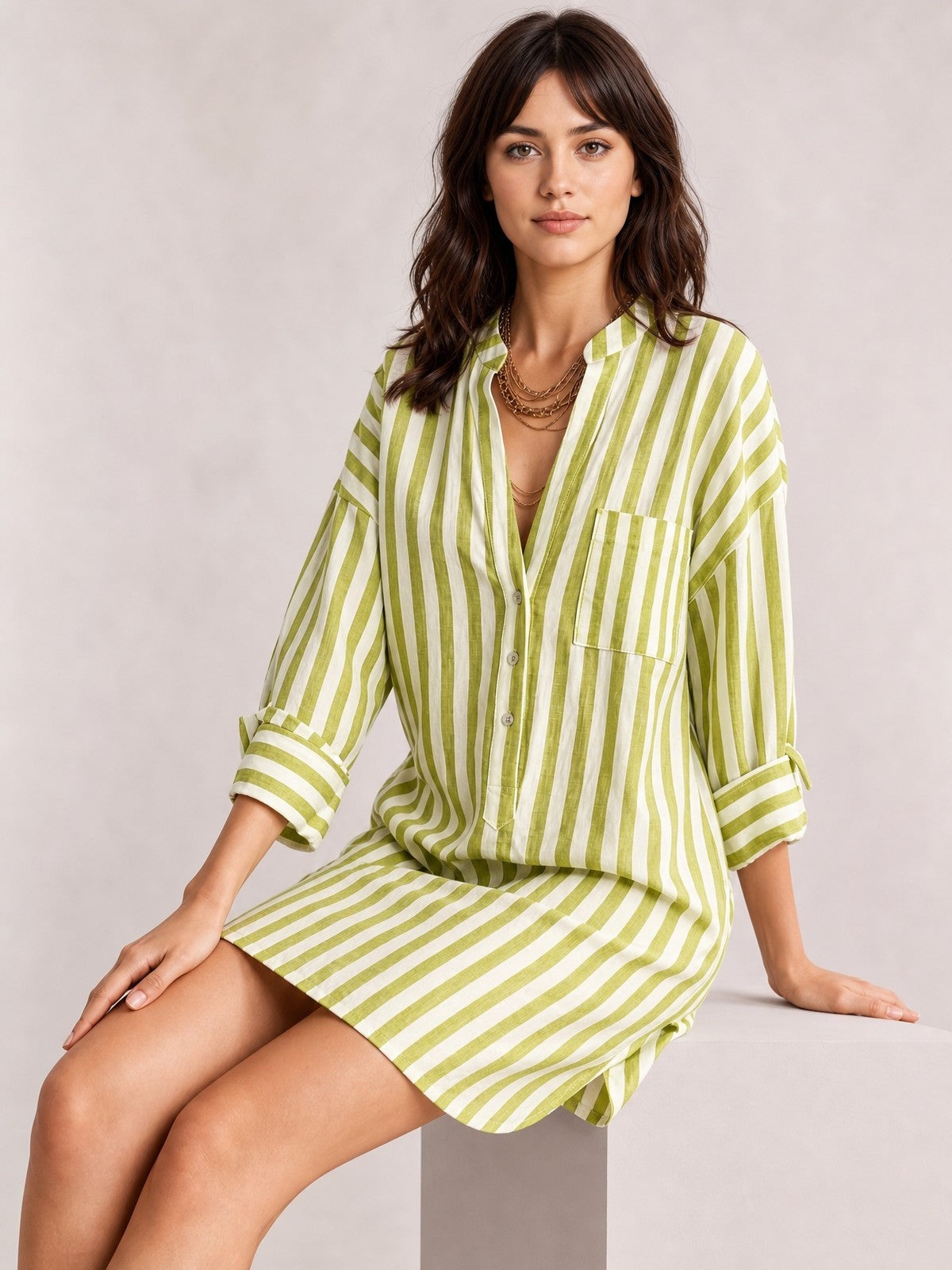 A seated model wears a green-and-white striped shirt mini dress with a relaxed silhouette, button front, chest pocket, long rolled sleeves, and above-the-knee hem.