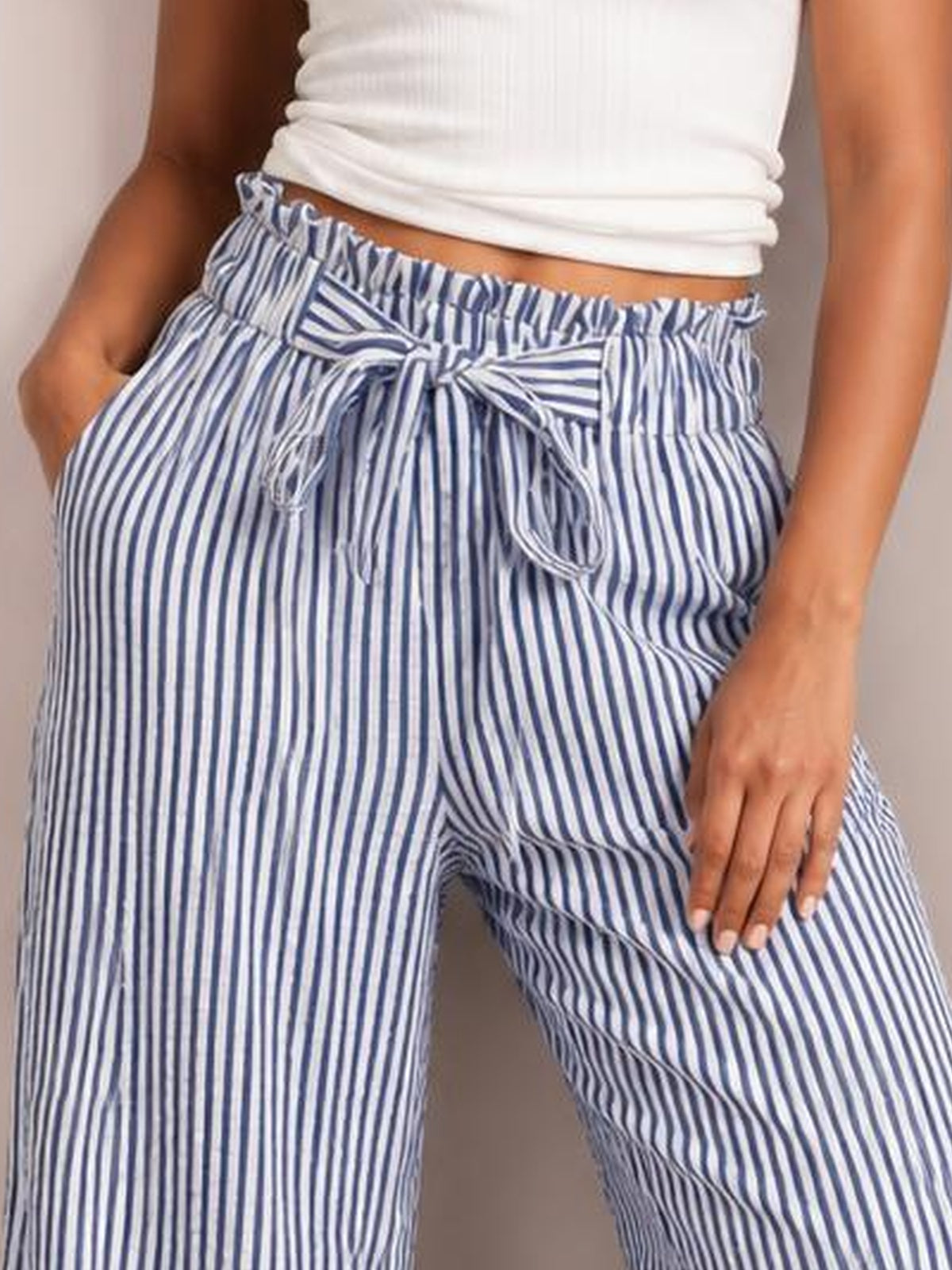 Model wears blue-and-white striped wide-leg paper bag pants with a gathered waist, front tie belt, and pockets, standing with one hand in a pocket.