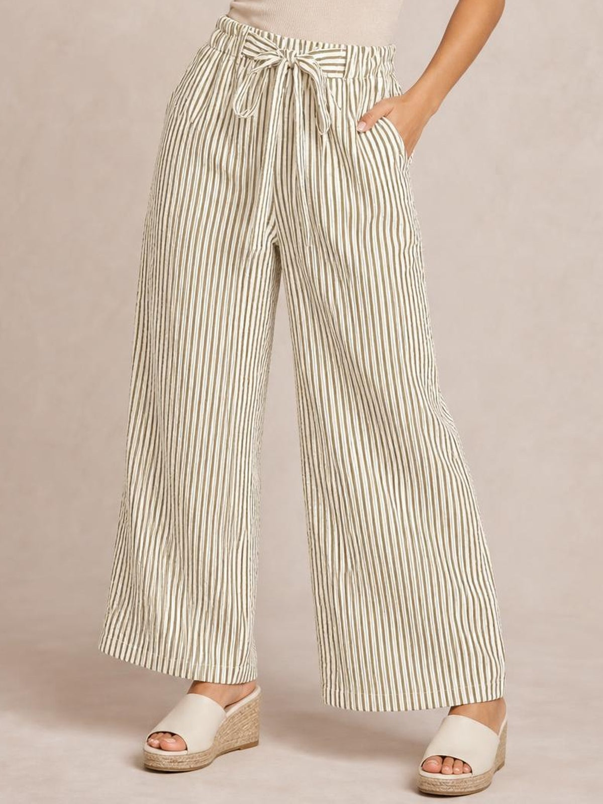 Model wears striped wide-leg paper bag pants in cream and taupe with a tied waist and side pockets.