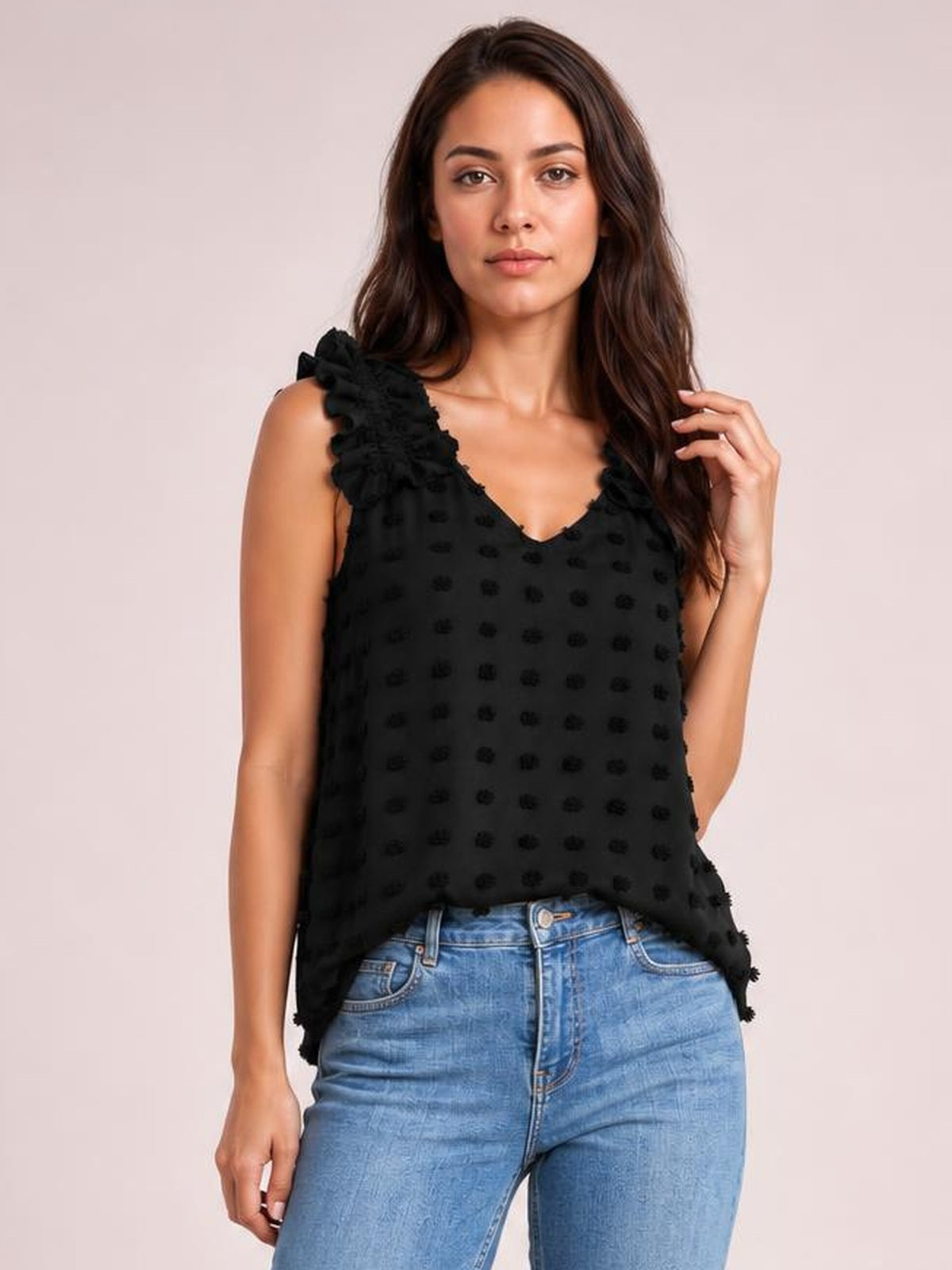 A model wears a black Swiss dot V-neck tank with ruffled shoulder straps and a relaxed, flowing silhouette, lightly touching her hair.
