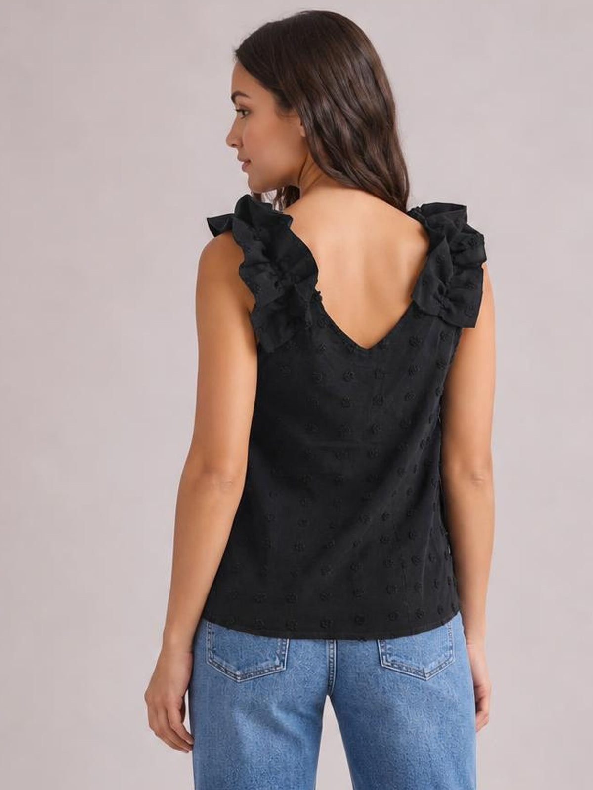 A model poses from the back in a black Swiss dot tank with a V-shaped back neckline, ruffled shoulder straps, and a straight, relaxed silhouette.