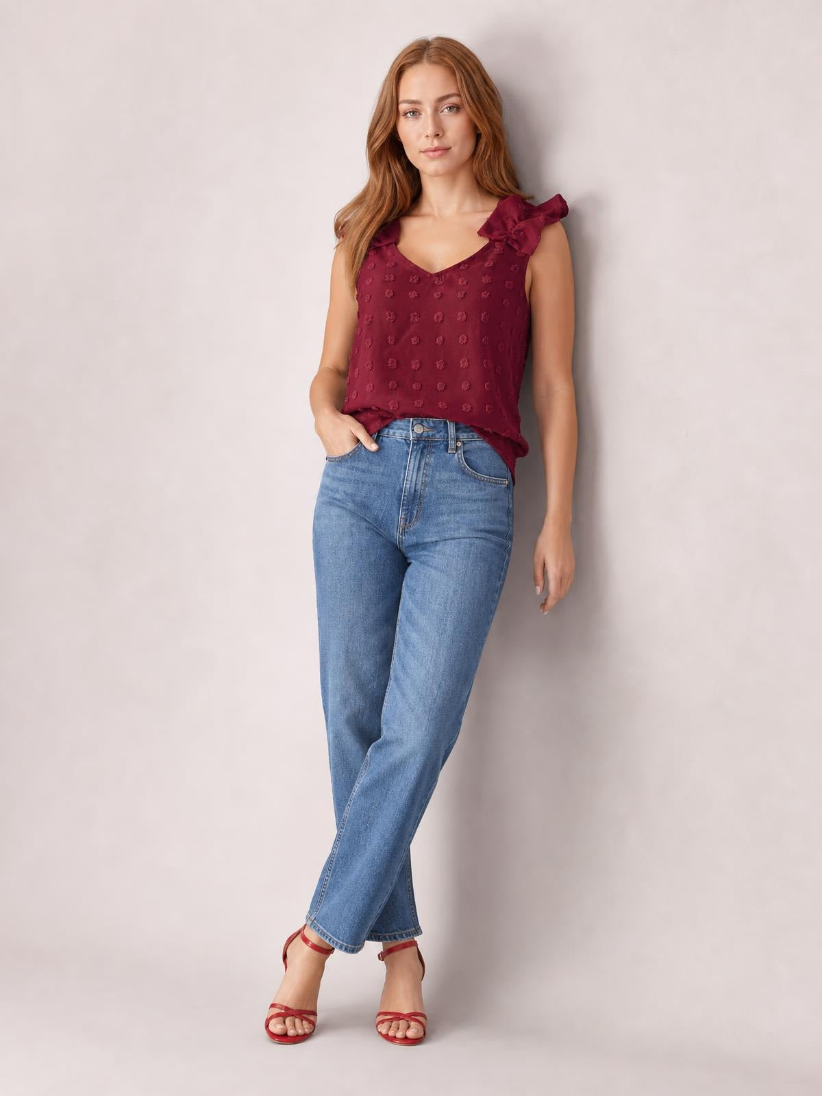 Model poses with one hand in her jeans pocket wearing a burgundy Swiss dot V-neck tank with a relaxed silhouette and ruffled shoulder straps.