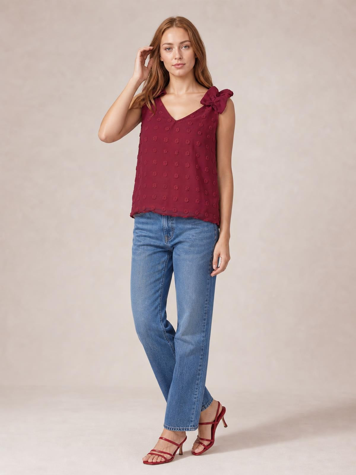 A model poses with one hand in her hair, wearing a burgundy Swiss dot V-neck tank with shoulder bows, a relaxed straight silhouette, and a straight hem.