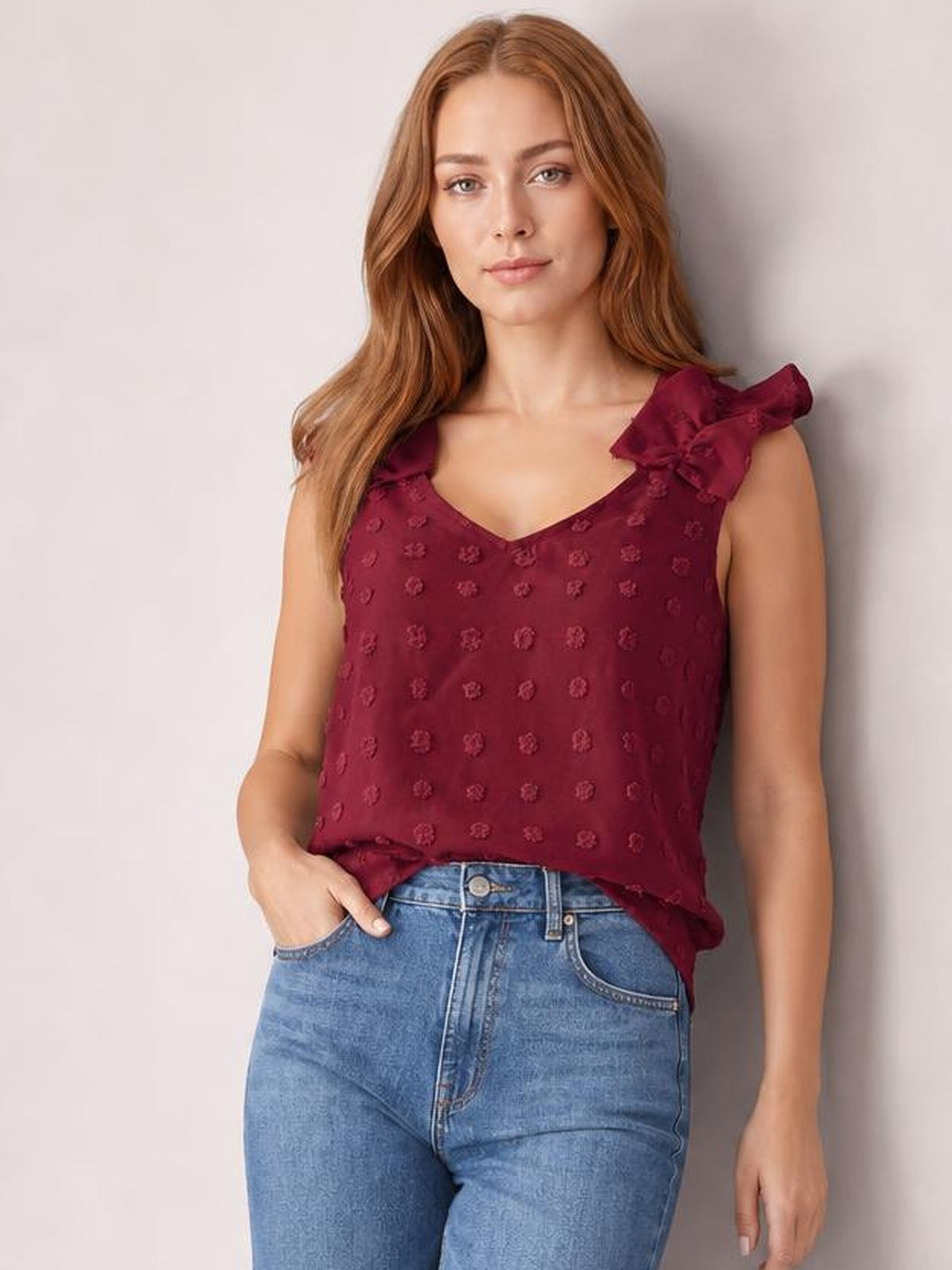 Model wears a burgundy Swiss dot V-neck tank with ruffled shoulder straps and a relaxed silhouette, posing with one hand in her jeans pocket.
