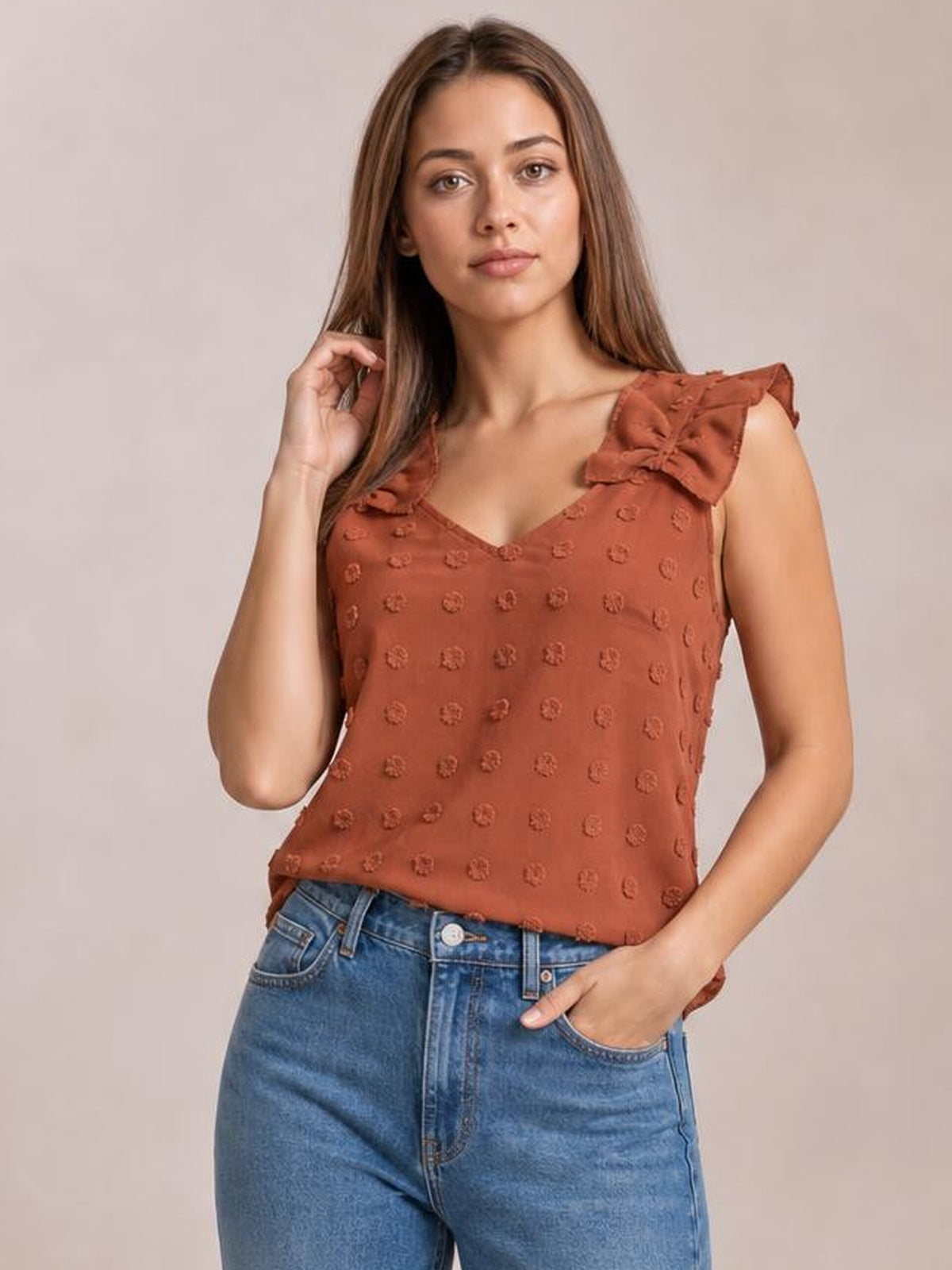 A model poses in a rust-colored Swiss dot V-neck tank with ruffled shoulder straps, one hand touching her hair and the other in her jeans pocket.