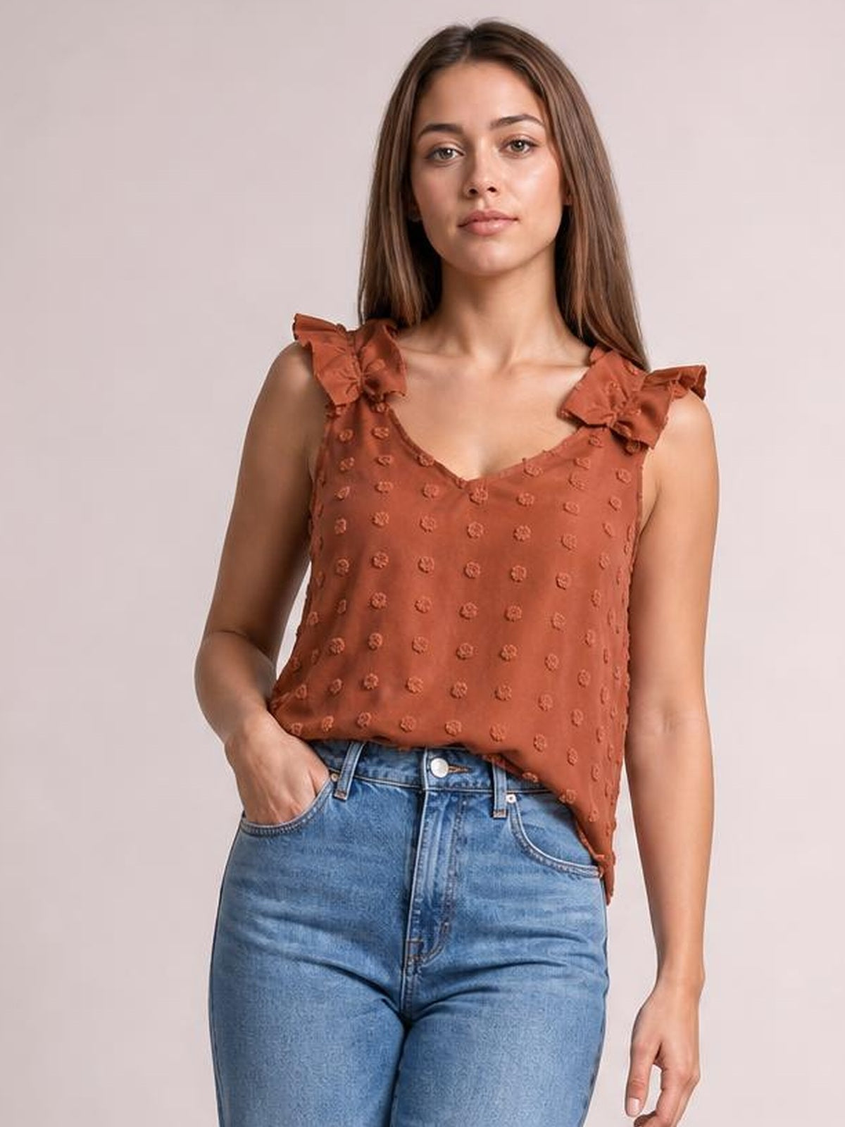 A model wears a rust Swiss dot V-neck tank with a relaxed silhouette, textured fabric, and ruffled shoulder straps, posing with one hand in her jeans pocket.