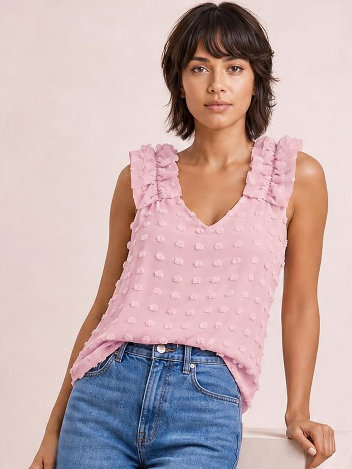 Model wears a pink Swiss dot V-neck tank with ruffled shoulder straps and a relaxed, slightly loose silhouette, leaning with one hand resting at her side.