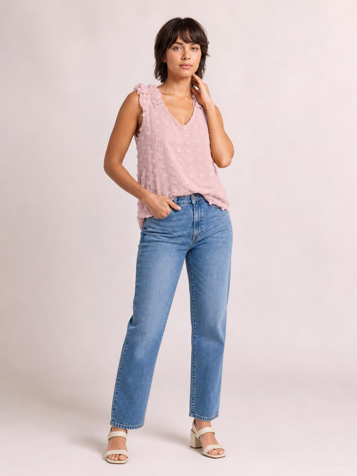 Model poses in a pink Swiss dot V-neck tank with ruffled shoulder straps and a relaxed straight silhouette, paired with light-wash jeans.