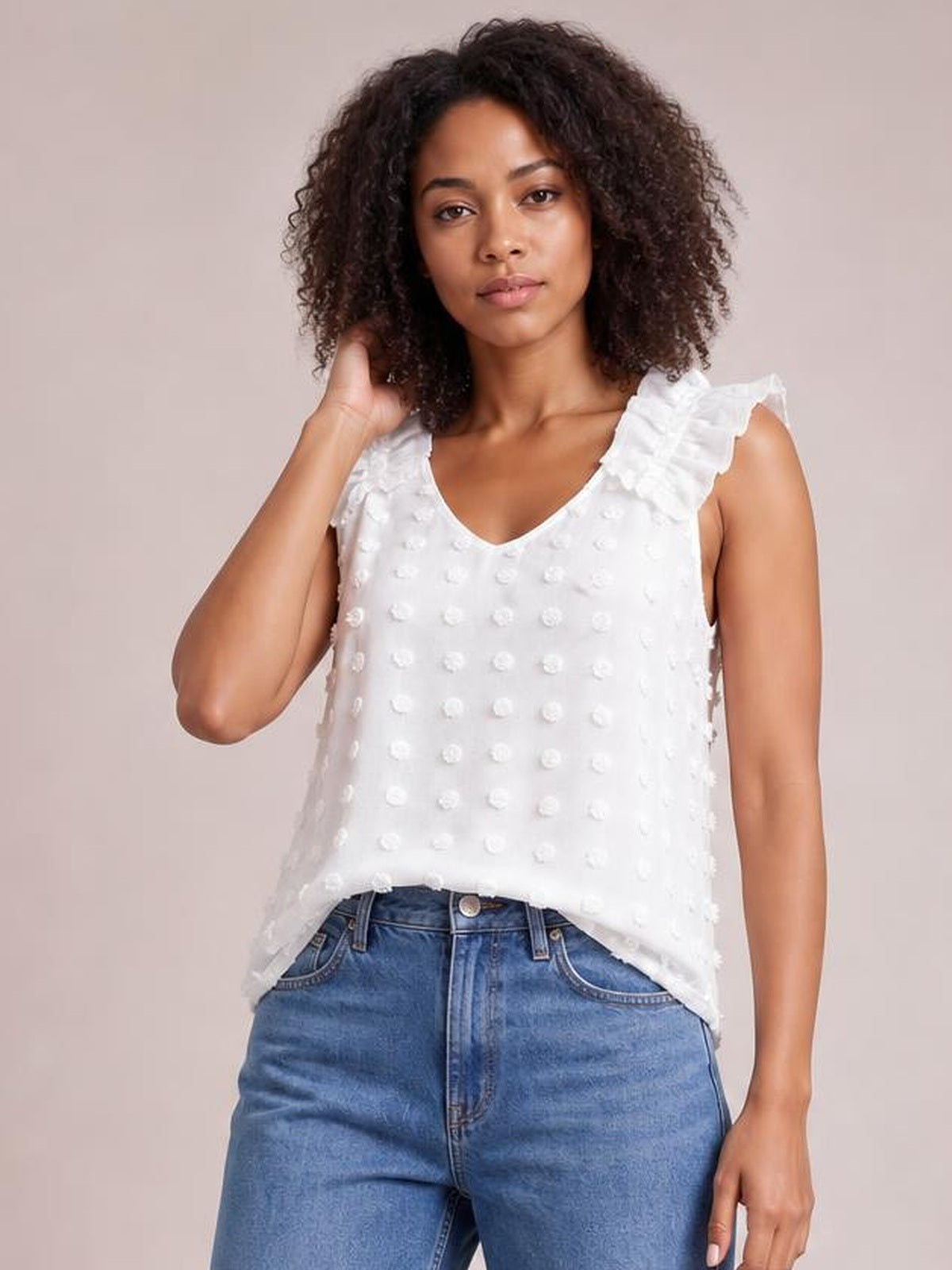 A model poses in a white Swiss dot V-neck tank with ruffled shoulder straps and a relaxed, straight silhouette, touching her hair with one hand.