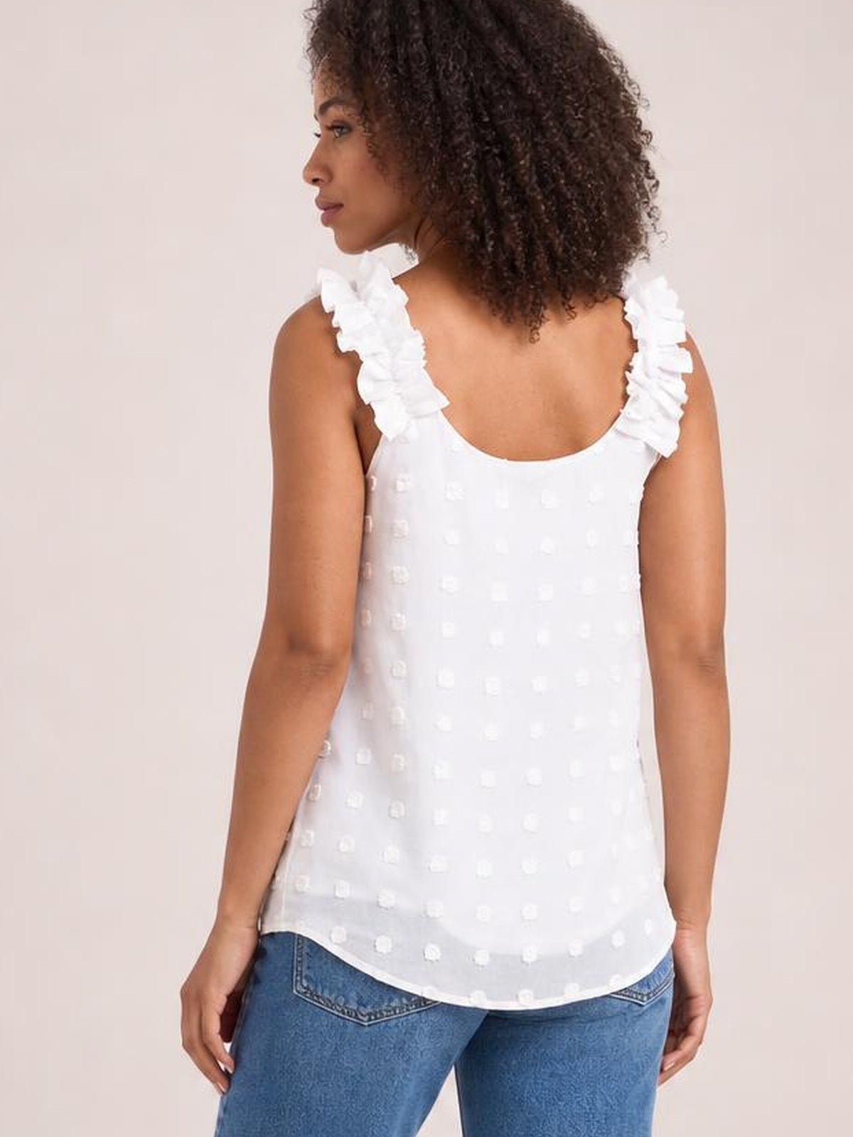 A model poses from the back in a white Swiss dot tank with ruffled shoulder straps, a gently curved hem, and a relaxed, straight silhouette.