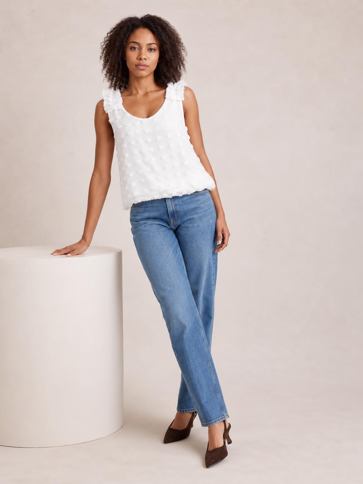 A model wears a white Swiss dot tank with a V-neck, ruffled shoulder straps, and a relaxed straight silhouette while leaning against a pedestal.