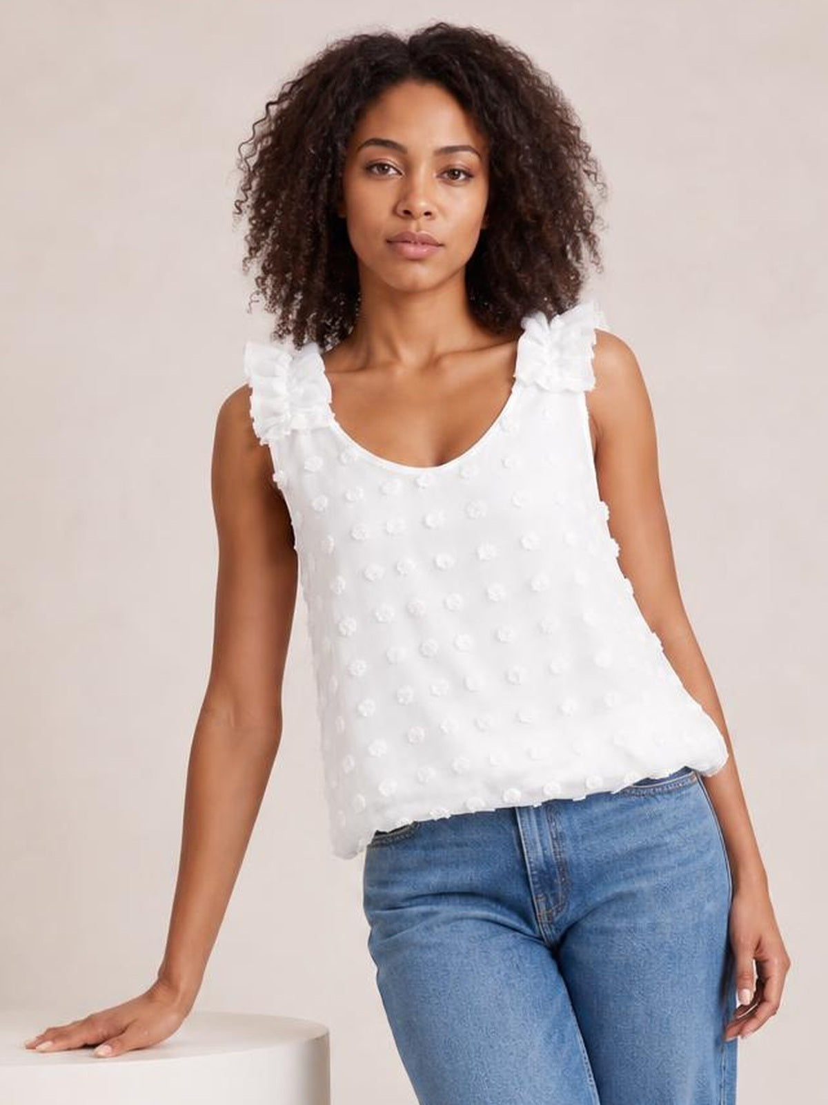 A model poses in a white Swiss dot tank with a scoop neckline, ruffled shoulder straps, and a relaxed, slightly flowing silhouette.