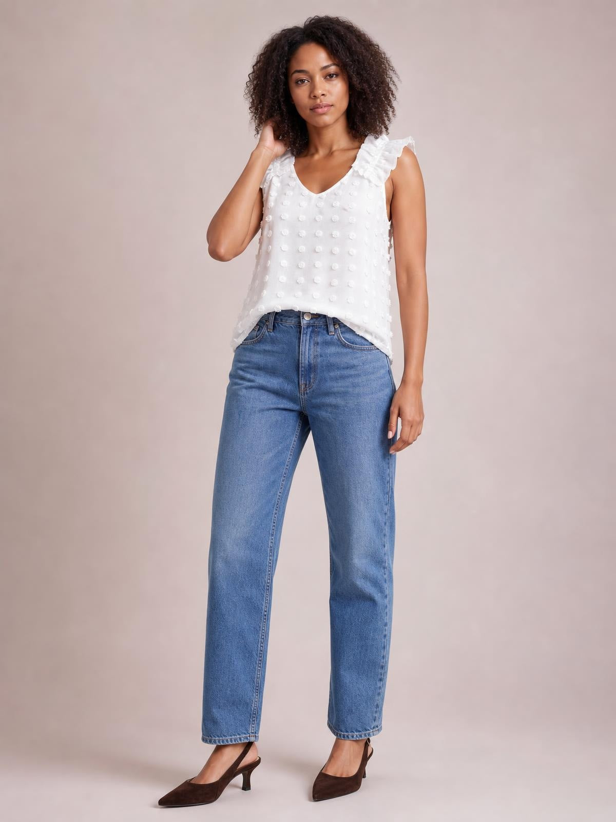 A model poses with one hand near her hair, wearing a white Swiss dot V-neck tank with ruffled shoulder straps and a relaxed straight silhouette.