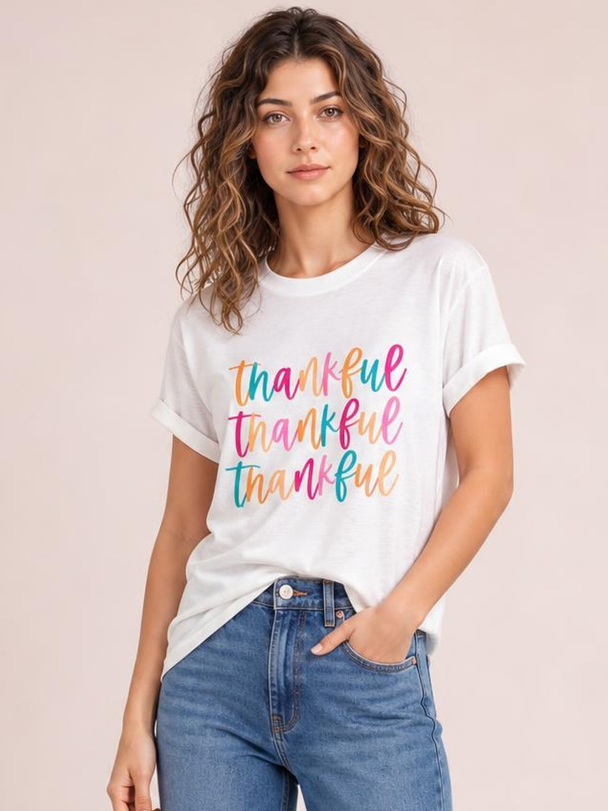 Model wearing a white THANKFUL round neck short sleeve T-shirt with rolled cuffs, colorful repeated lettering, and a relaxed silhouette, posing with one hand in her jeans pocket.