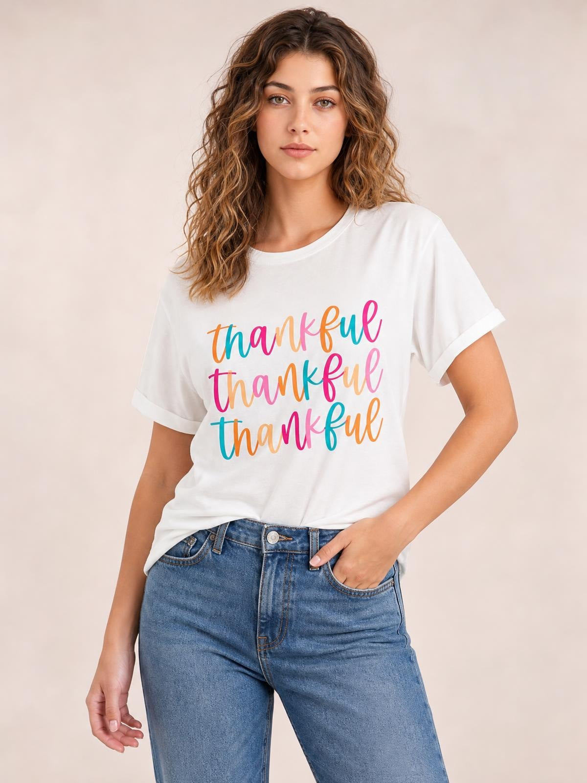 Model wearing a white THANKFUL round neck short sleeve T-shirt with colorful repeated text, rolled sleeve cuffs, and a relaxed silhouette, posing with one hand in her jeans pocket.