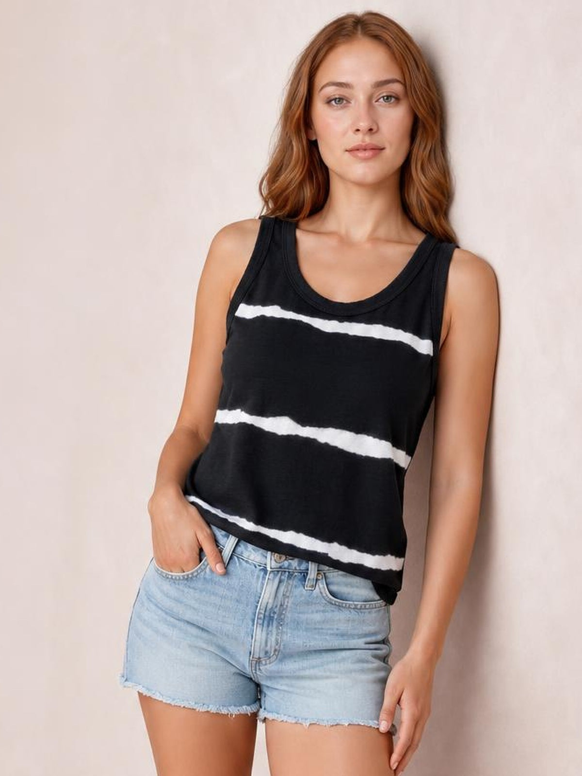 Model wearing a black tie-dye round neck tank with white horizontal bands, wide shoulder straps, and a relaxed silhouette, posing with one hand in her shorts pocket.