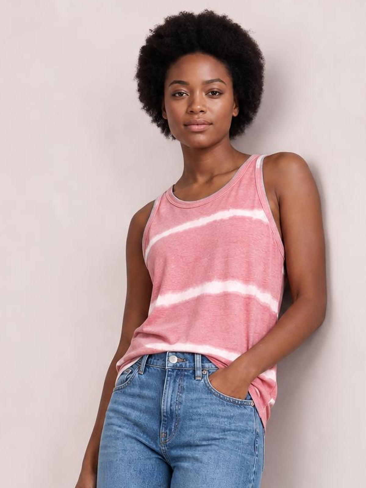 A model wears a pink tie-dye round neck tank with white horizontal bands, sleeveless construction, and a relaxed silhouette, posing with one hand in her jeans pocket.