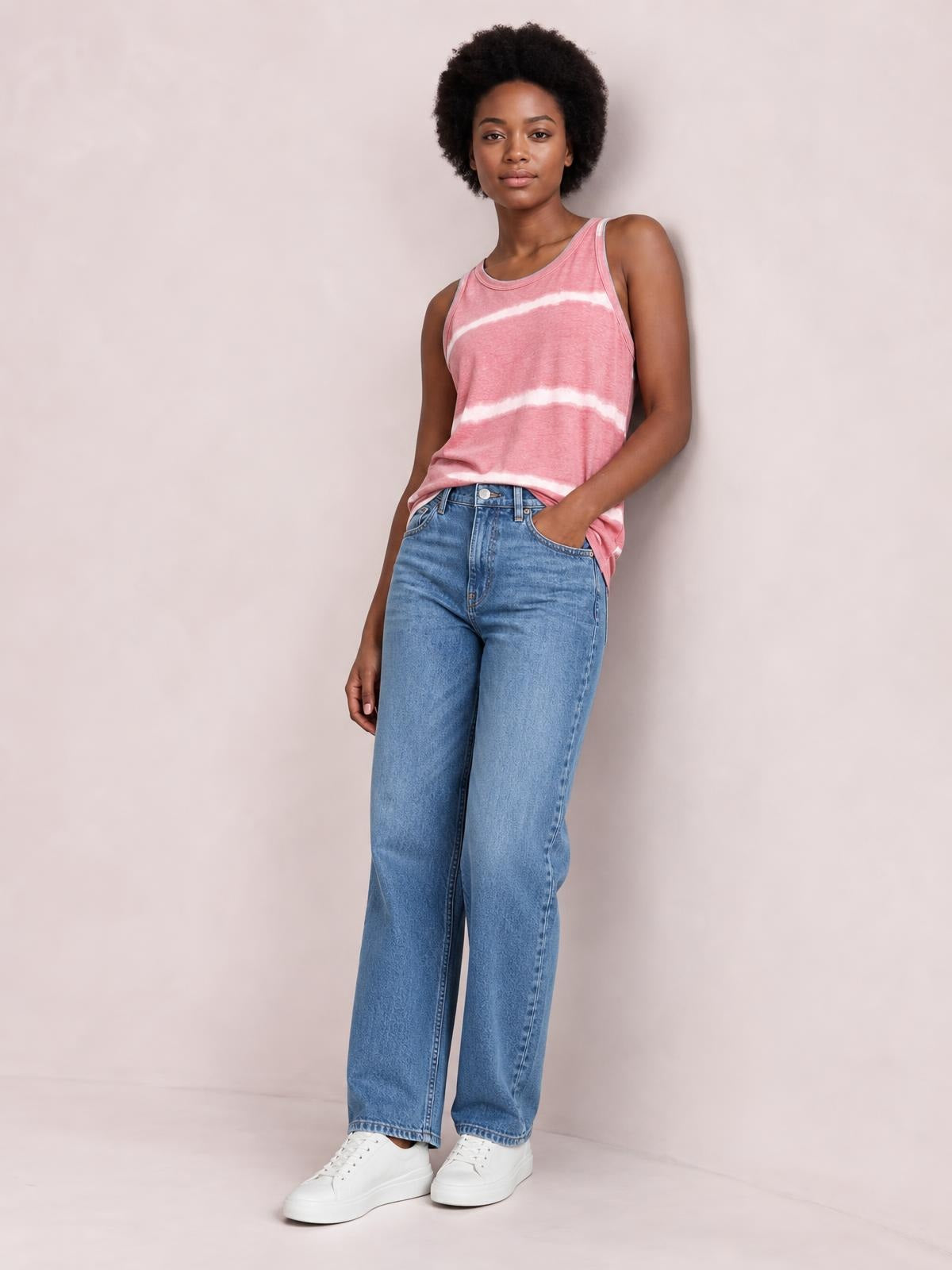 Model wears a pink and white tie-dye round neck tank with a relaxed straight silhouette and wide shoulder straps, posing with one hand in her jeans pocket.
