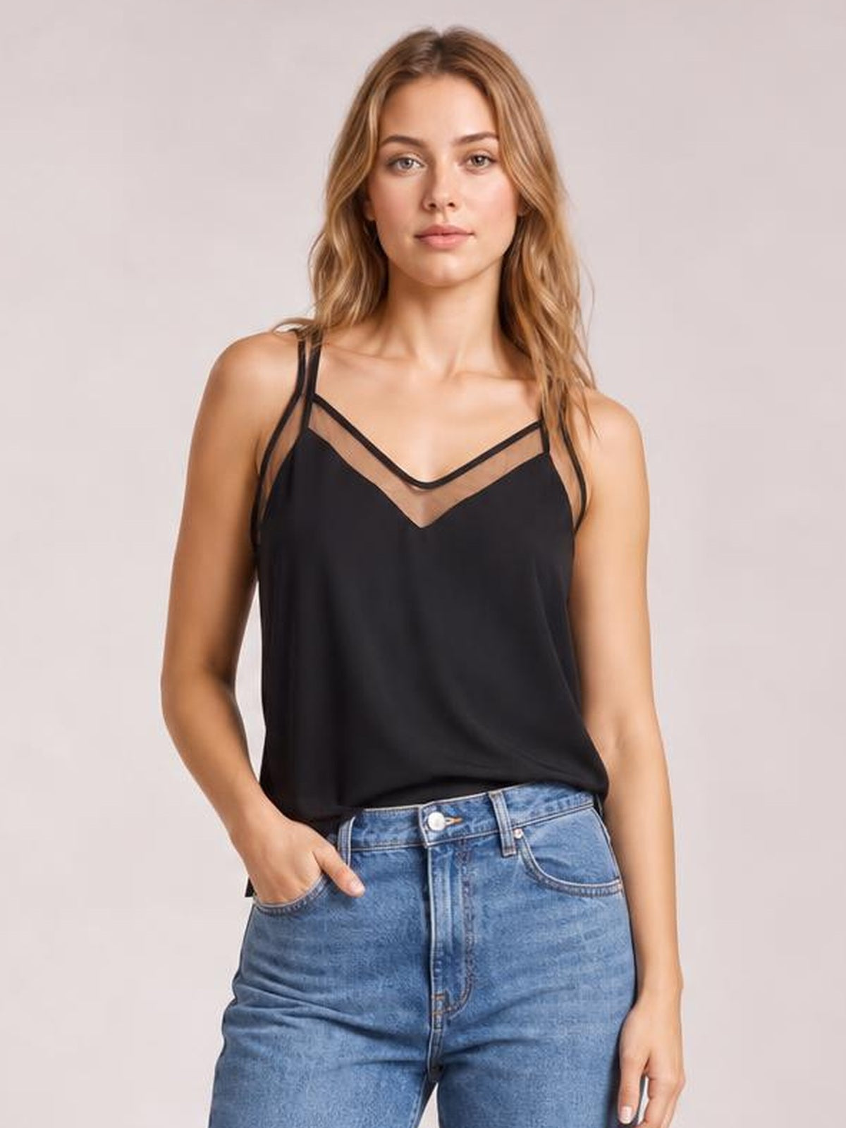 A model wears a black V-neck spaghetti strap cami with a semi-sheer neckline, relaxed silhouette, and straight construction while standing with one hand in her jeans pocket.