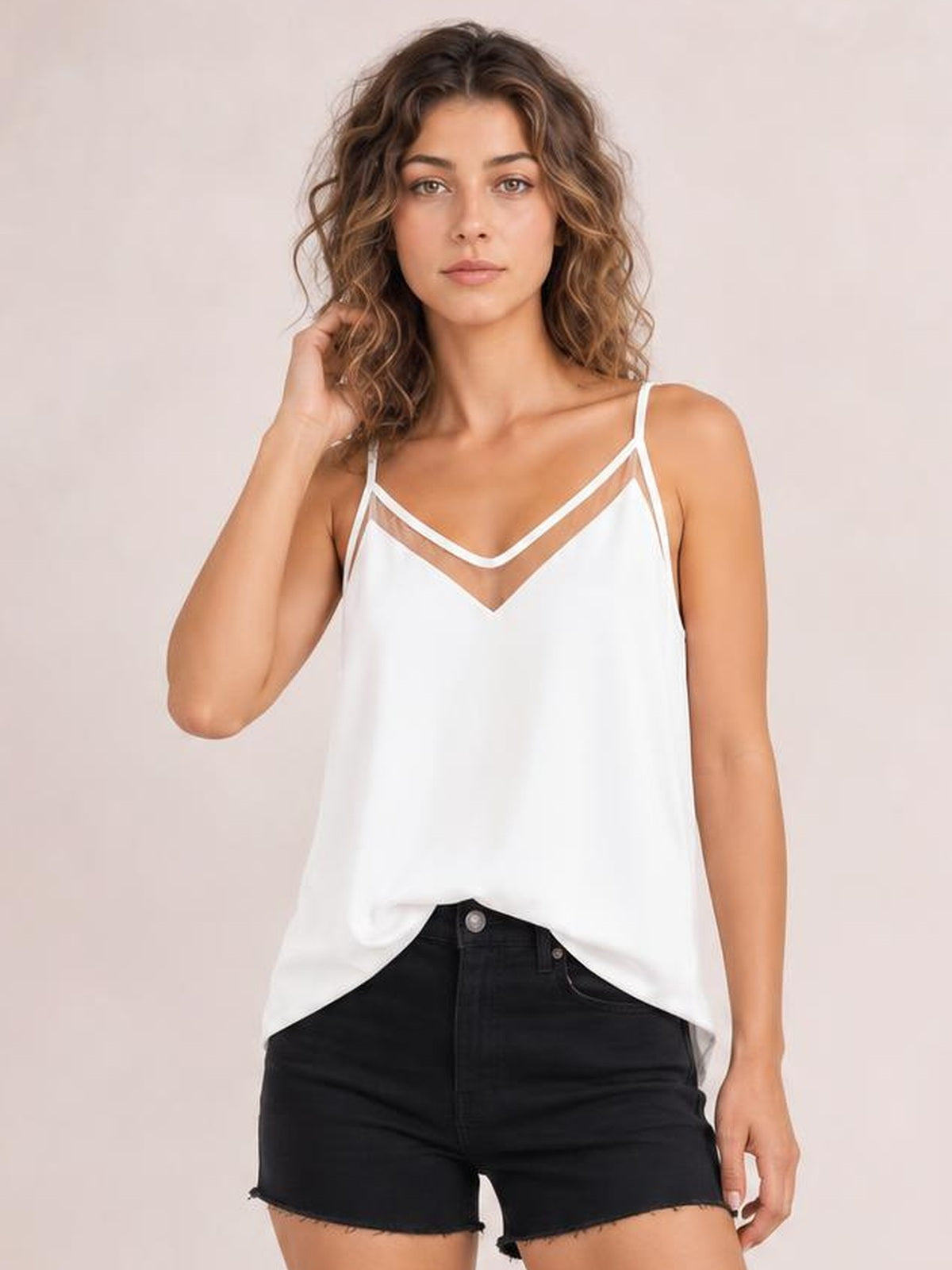 Model wearing a white V-neck spaghetti strap cami with semi-sheer layered neckline, relaxed straight silhouette, and curved hem, posing with one hand in her hair.