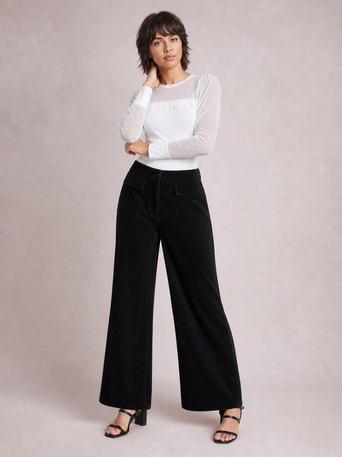 A model poses with one hand at her neck, wearing black wide-leg corduroy pants with a drawstring waistband and roomy patch pockets.