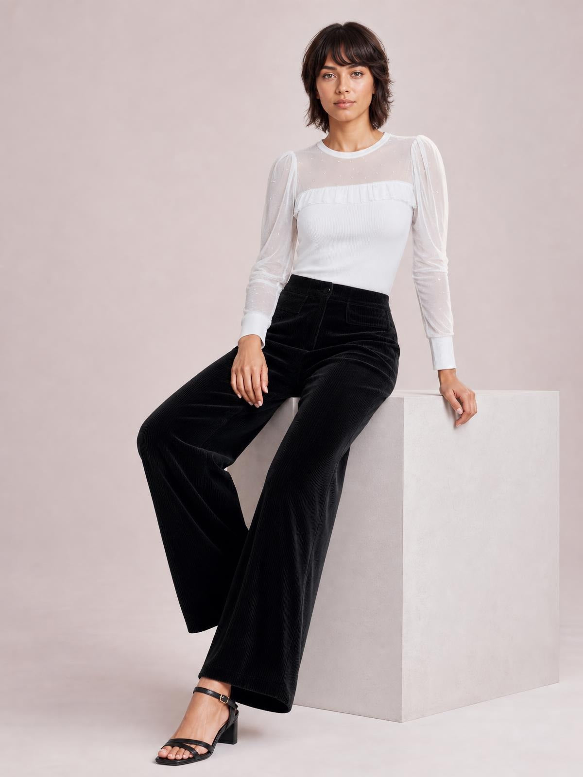 A seated model wears black wide-leg corduroy pants with a high-rise waistband, button closure, pocketed construction, and flowing full-length legs.