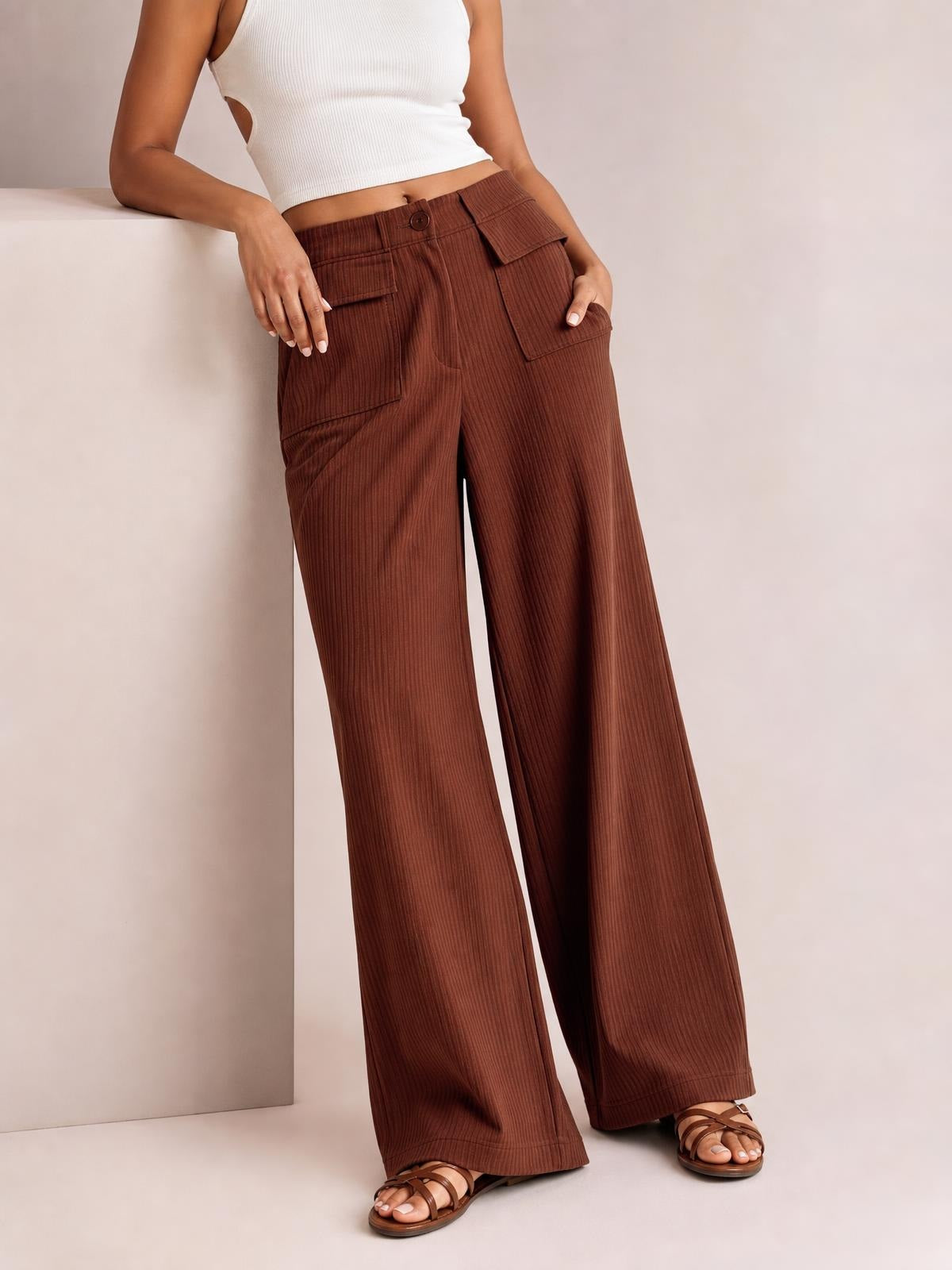A model leans against a ledge wearing brown wide-leg corduroy pants with a buttoned waistband, vertical ribbed texture, and roomy front flap pockets.