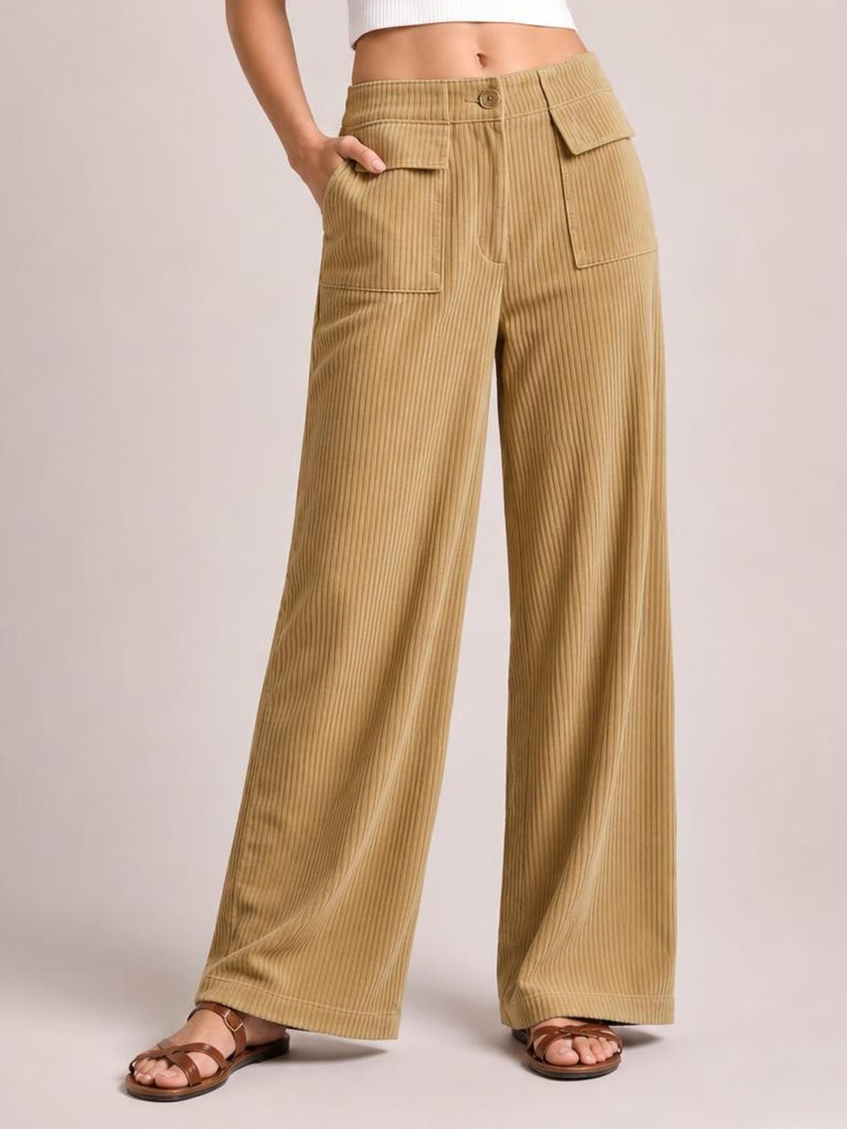 Model wears tan wide-leg corduroy pants with a high-rise button closure, vertical ribbed texture, and large flap pockets, standing with one hand in a pocket.