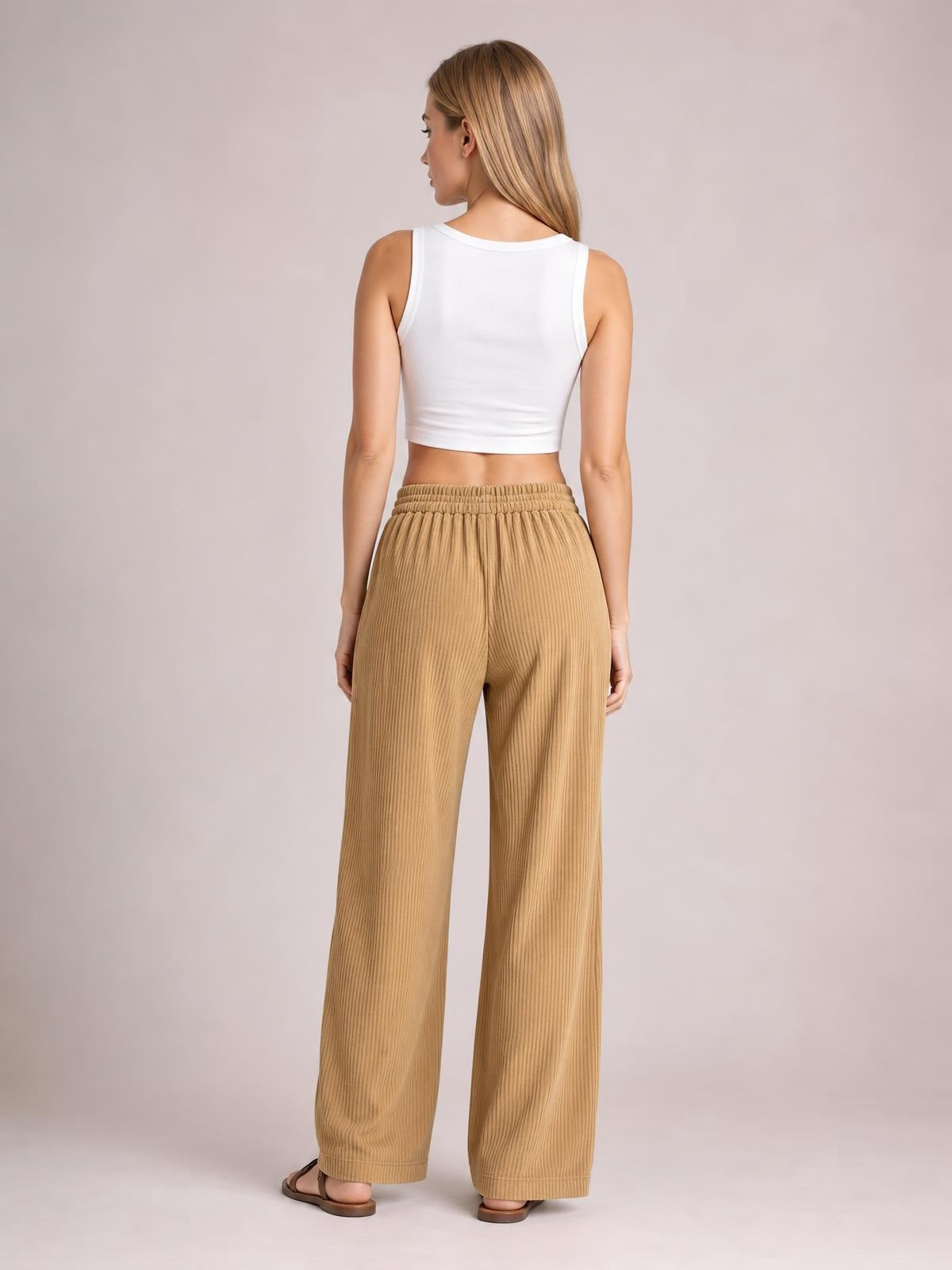 A model stands facing away in tan wide-leg corduroy pants with a relaxed silhouette, ribbed texture, elastic waistband, and full-length legs.