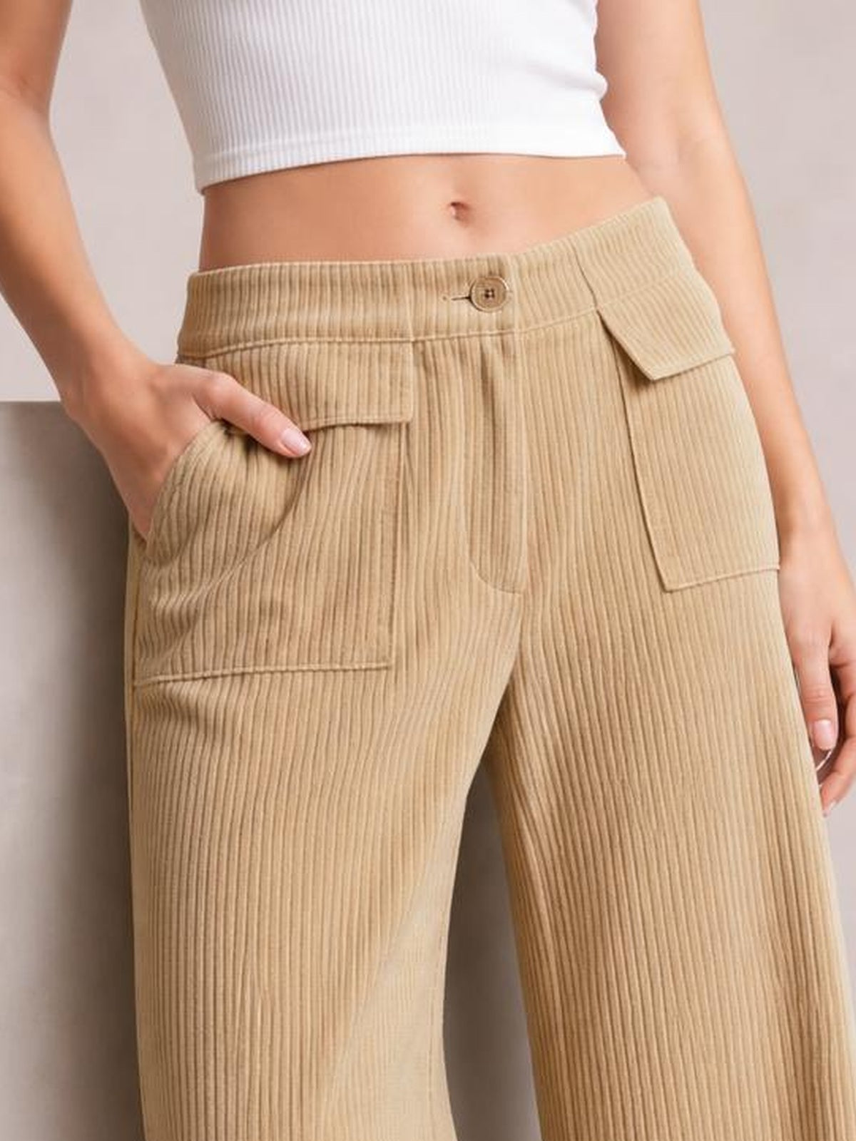 Model wears beige wide-leg corduroy pants with a button waistband, vertical ribbed texture, and multiple flap pockets, posing with one hand in a pocket.