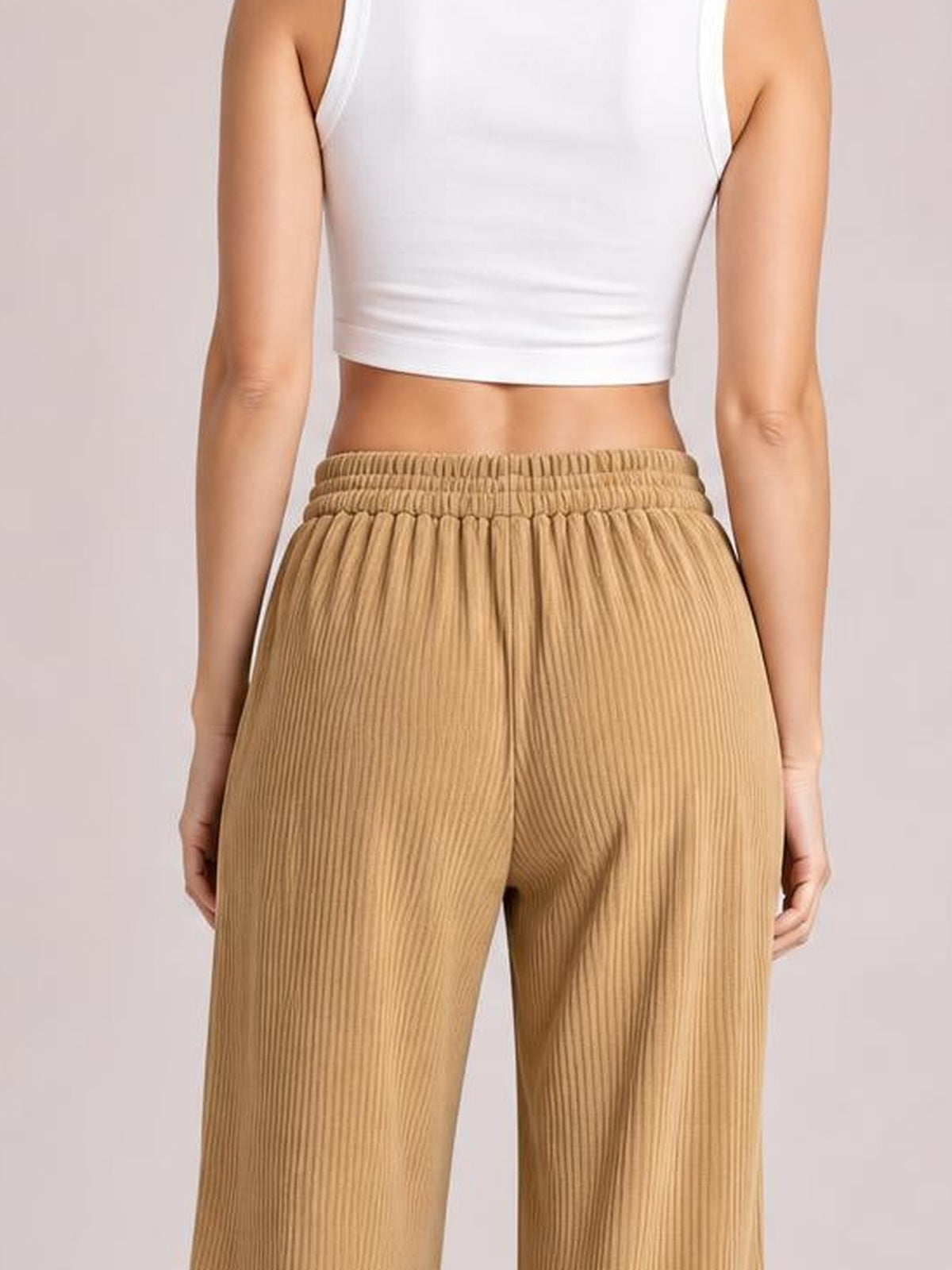 A model stands with hands at her sides, showing tan wide-leg corduroy pants with vertical ribbing, a gathered elastic waistband, and a relaxed silhouette.