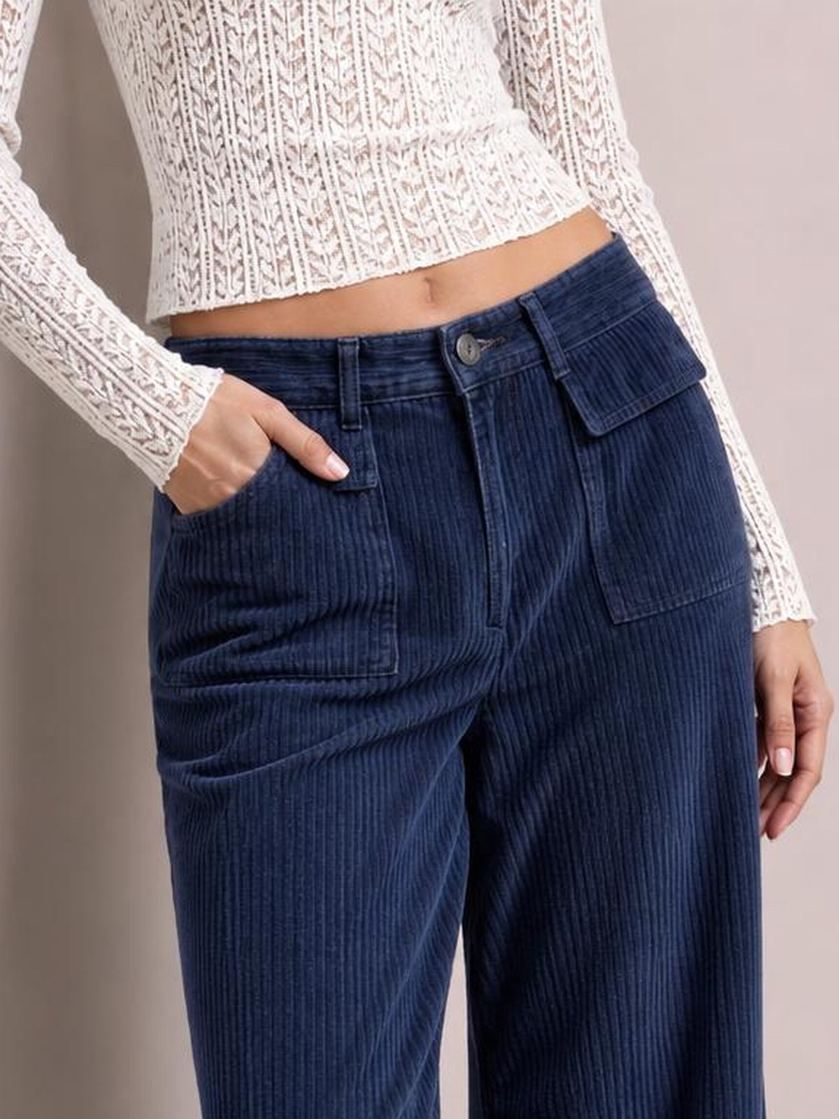 A model wears navy blue wide-leg corduroy pants with vertical ribbing, belt loops, a button closure, and multiple patch pockets, one hand tucked inside.