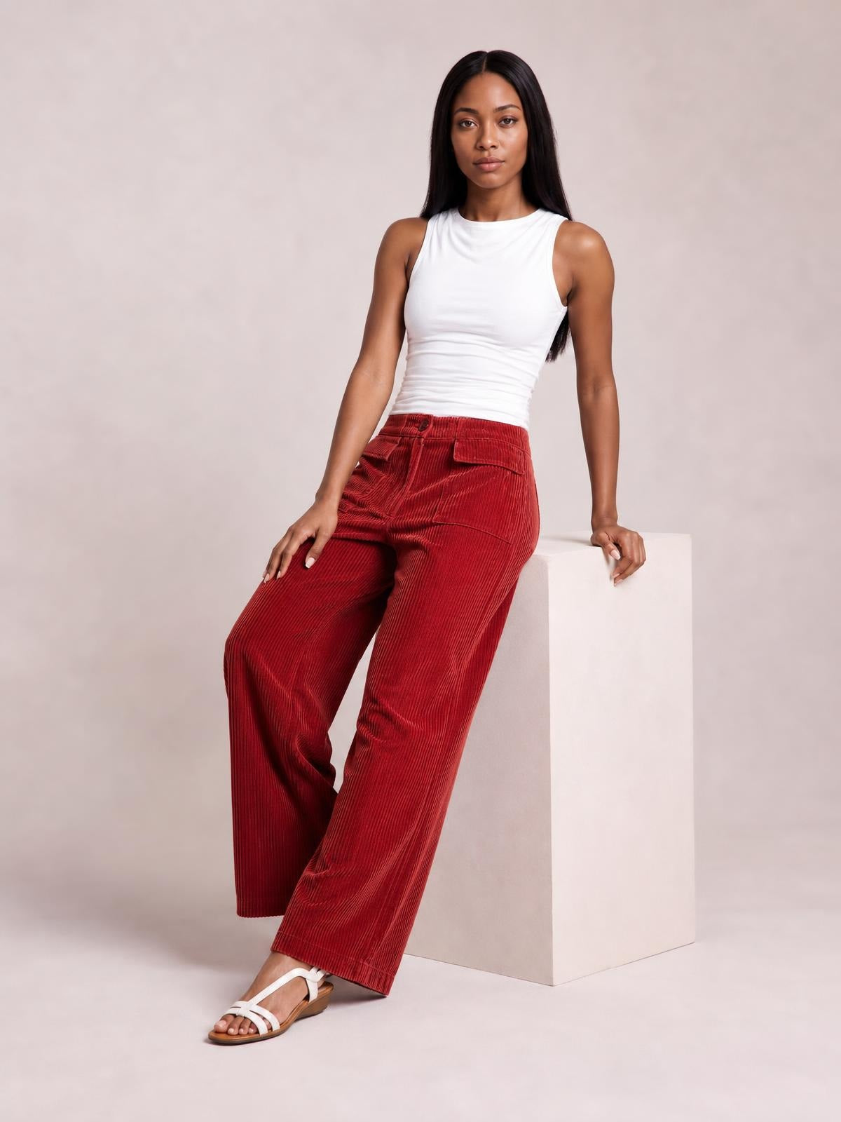 A model sits in red corduroy wide-leg pants with ribbed texture, front pockets, and a relaxed full-length silhouette, posing with one leg crossed.