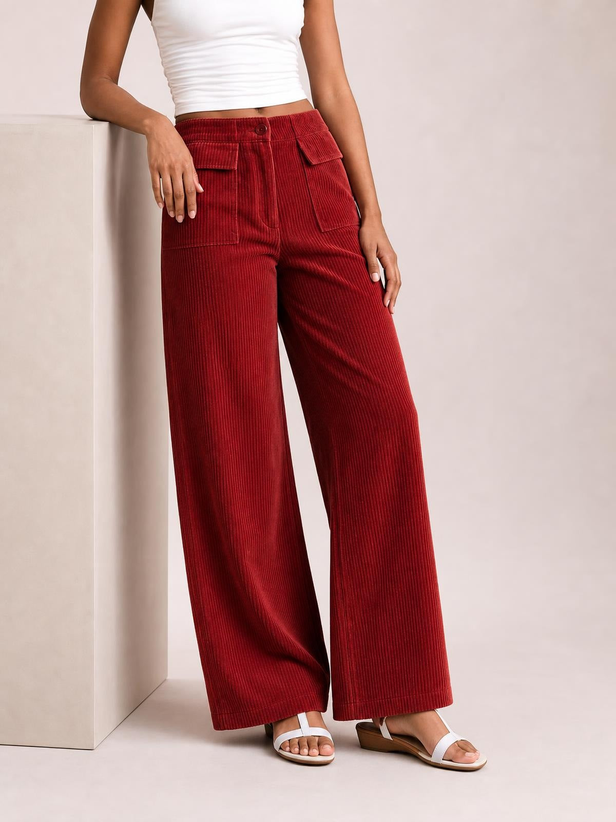 Model in a relaxed pose wears red wide-leg corduroy pants with a button waist, vertical ribbed texture, and multiple front pockets.