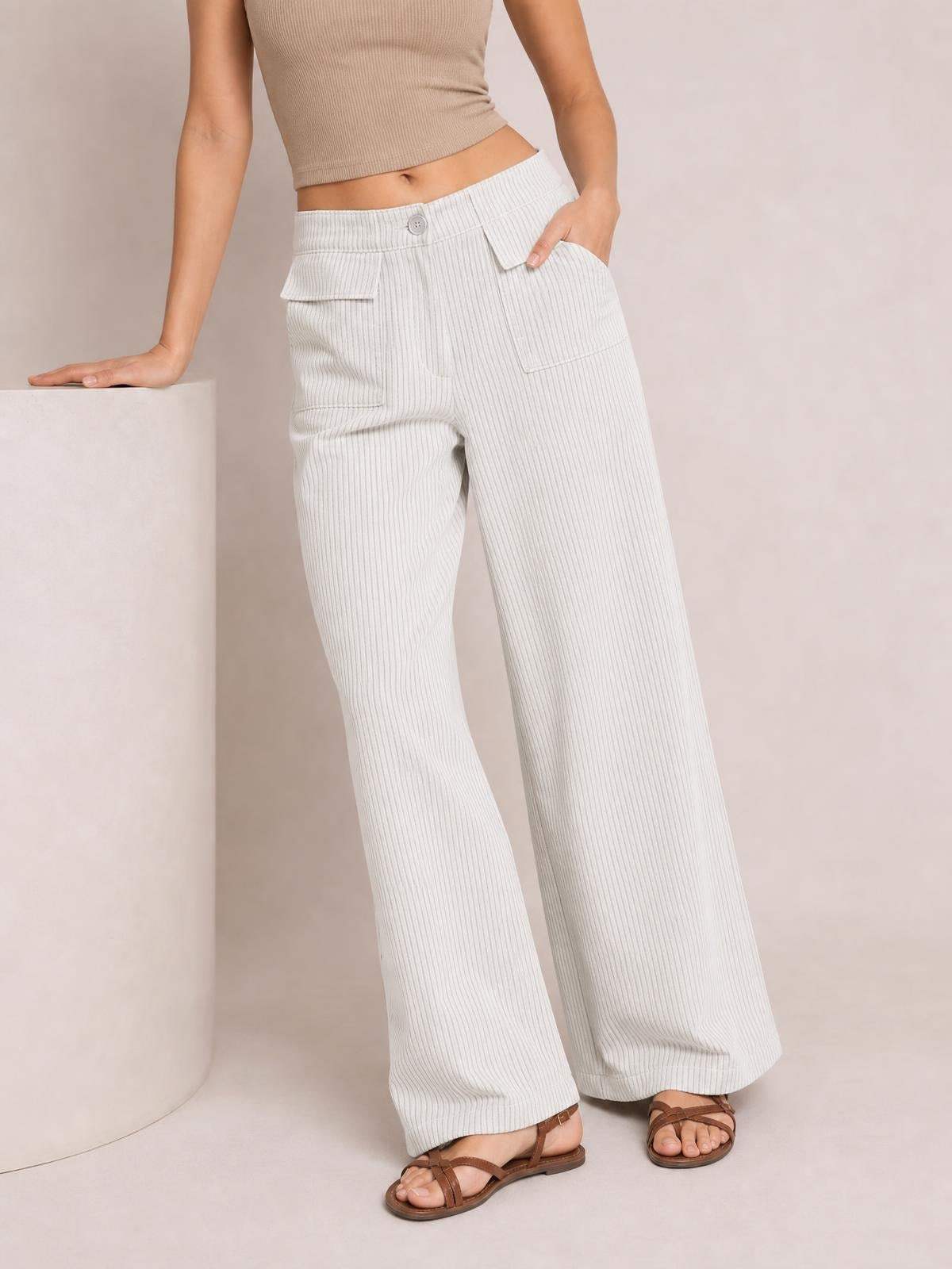 A model wears light gray wide-leg corduroy pants with a button waistband, vertical rib texture, patch pockets, and a relaxed silhouette while leaning and posing.