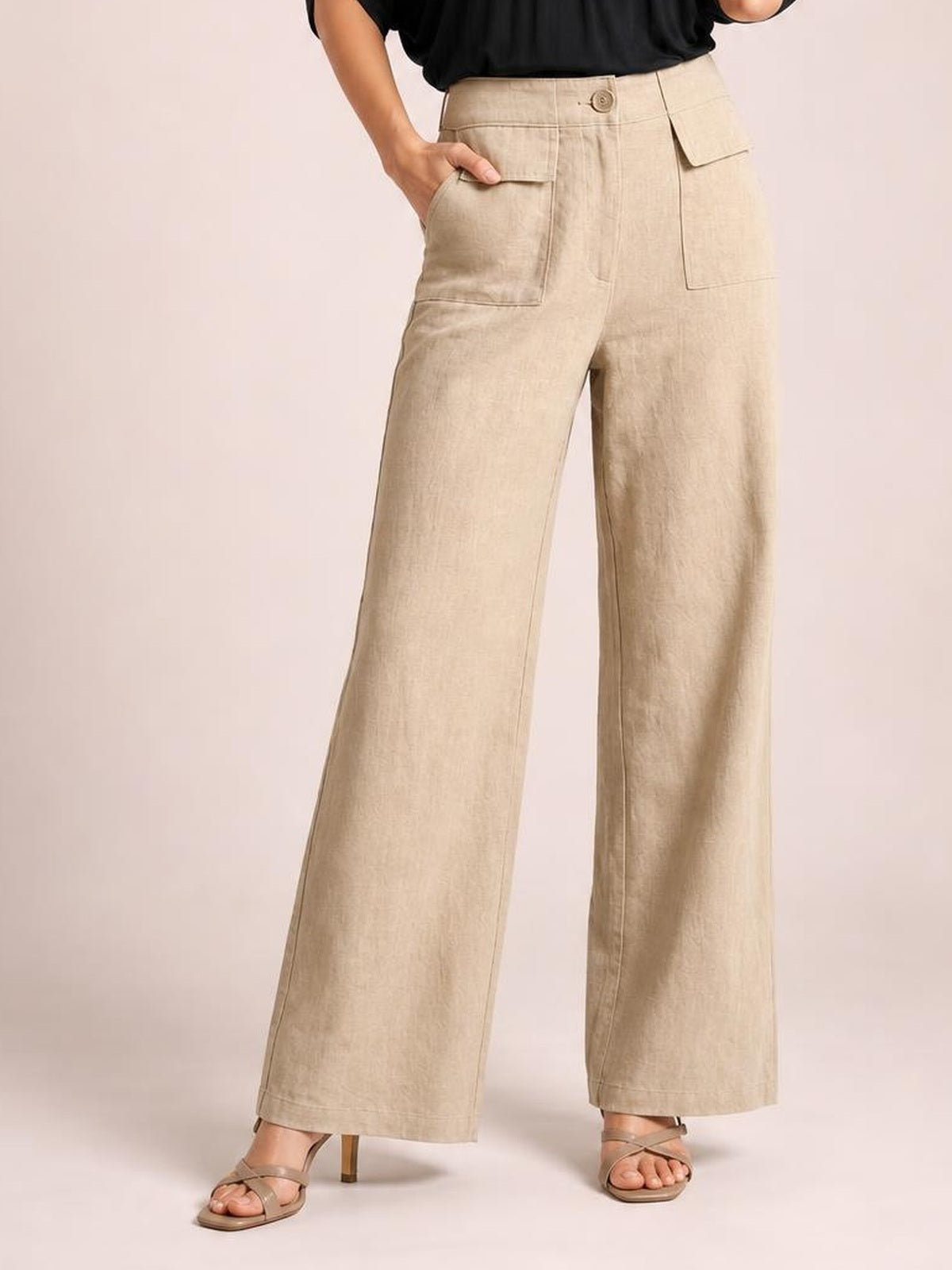 Model wearing beige high-waisted wide leg pants with a button closure, stitched paneling, and large patch pockets, posing with one hand tucked into a pocket.