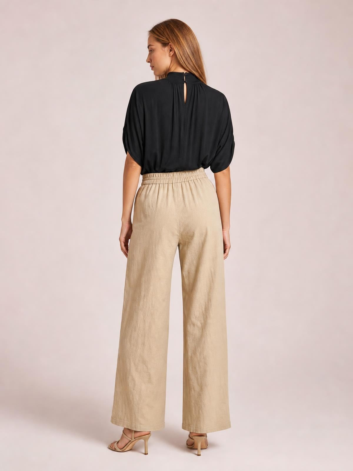 A model stands facing away in beige wide-leg pants with an elasticized waistband, relaxed straight silhouette, and ankle-length hems.