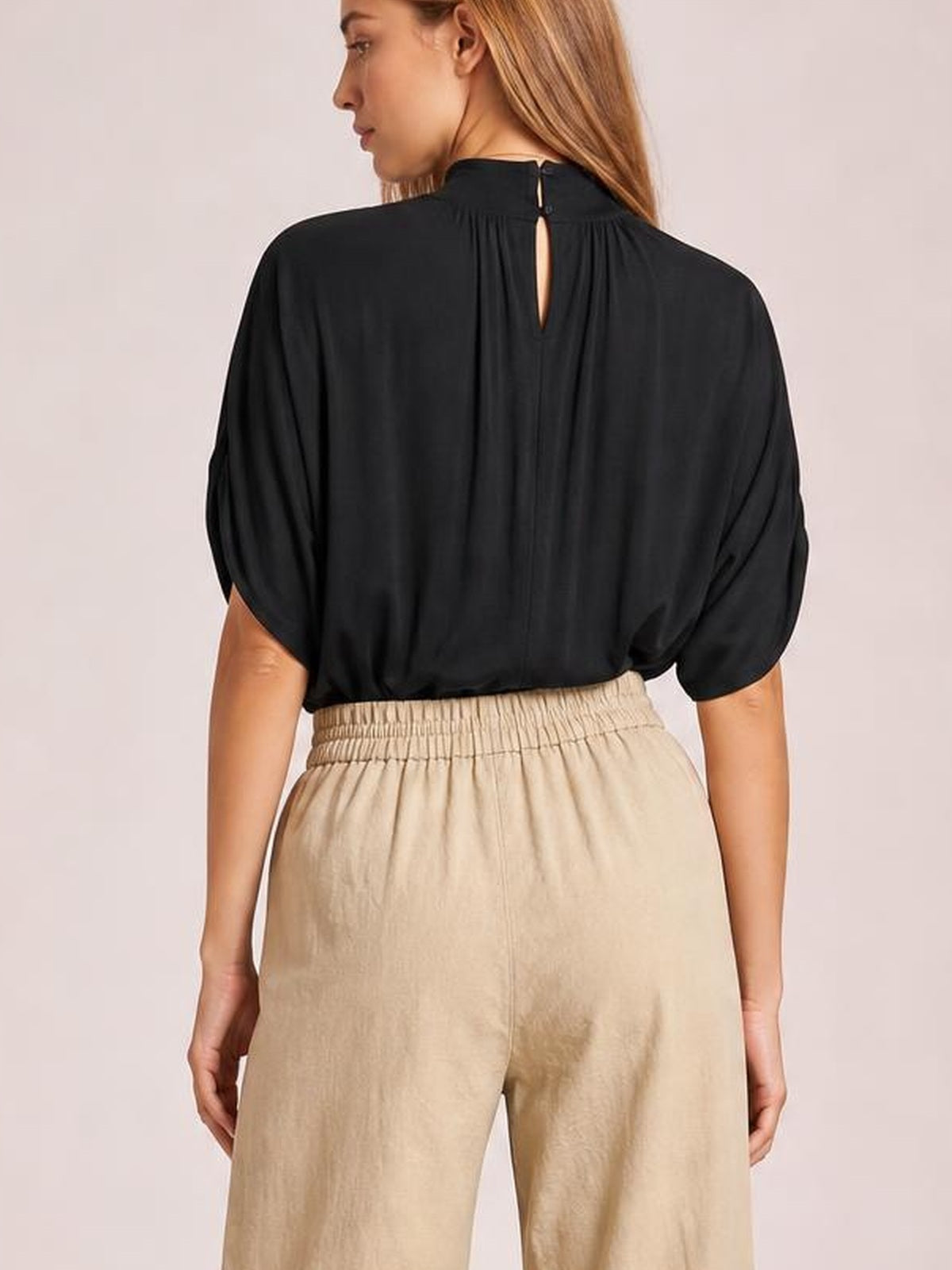 Model shown from the back wearing beige wide-leg pants with a relaxed silhouette and gathered elastic waistband, paired with a black short-sleeve top.