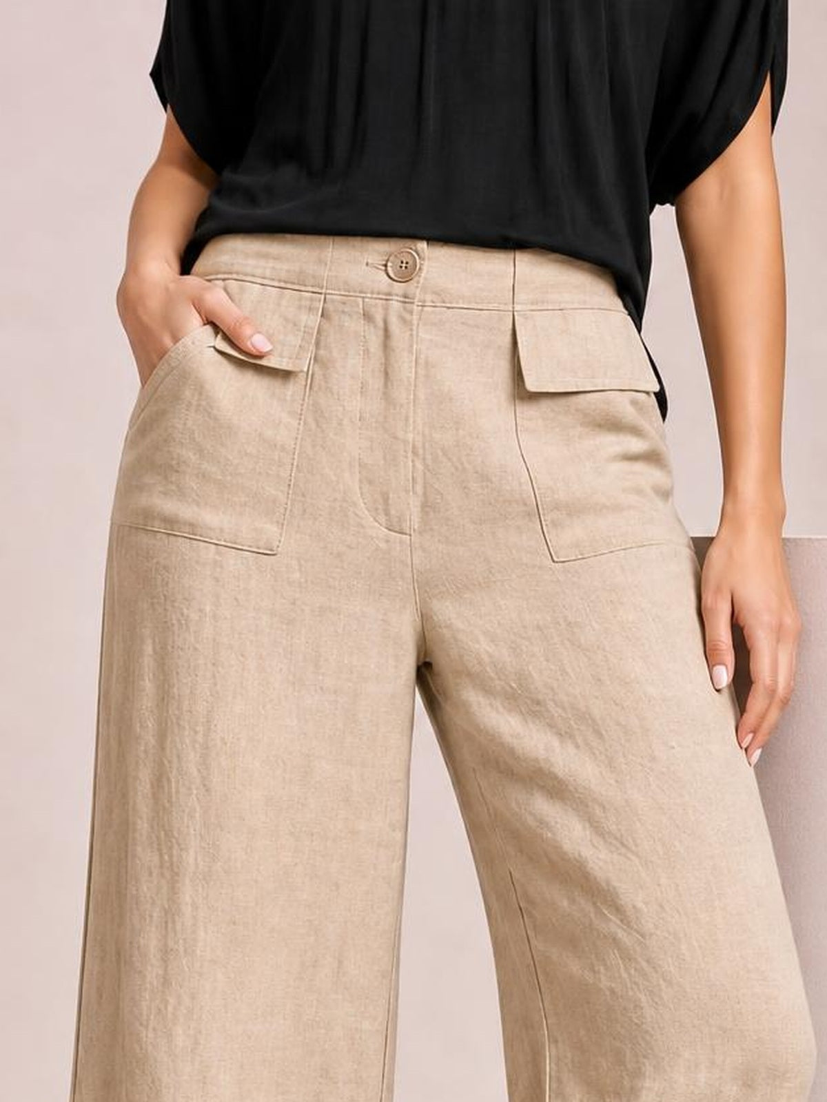 A model wears beige wide-leg pants with a structured waistband, button closure, stitched front patch pockets, and one hand resting in a pocket.