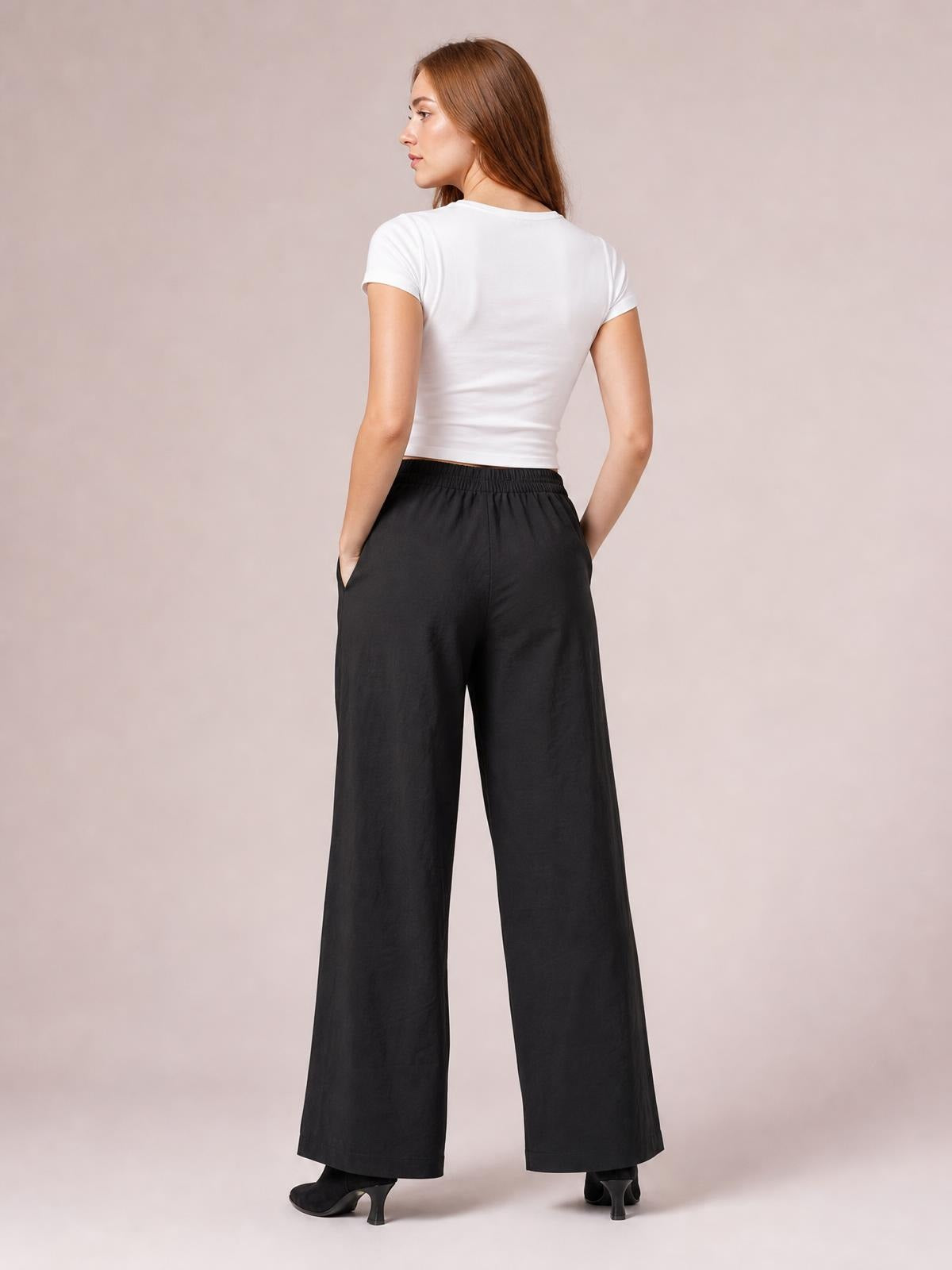 A model stands facing away in black wide leg pants with a gathered elastic waistband, side pockets, and a long, relaxed silhouette.